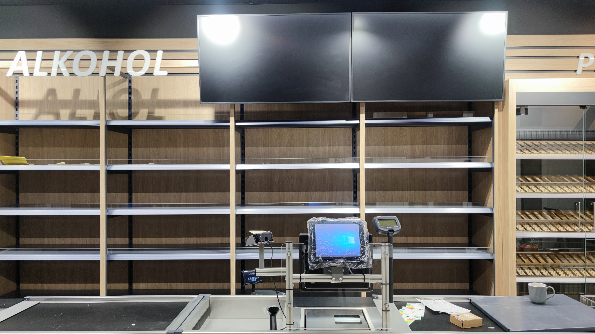 Puste półki sklepowe podświetlone od góry, z napisem 'ALKOHOL' i stanowiskiem kasowym z ekranem w folii ochronnej, sugerujące remont lub modernizację sklepu.