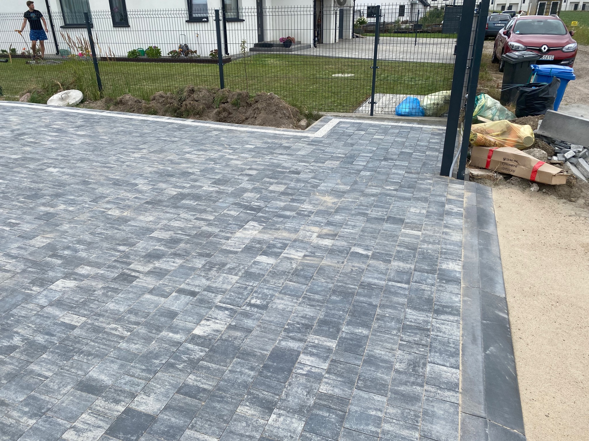 Ułożona szara kostka brukowa na podjeździe, obrzeża z ciemnej kostki, w tle dom z białymi ścianami i czarnymi oknami, widoczne worki z materiałami budowlanymi i samochód
