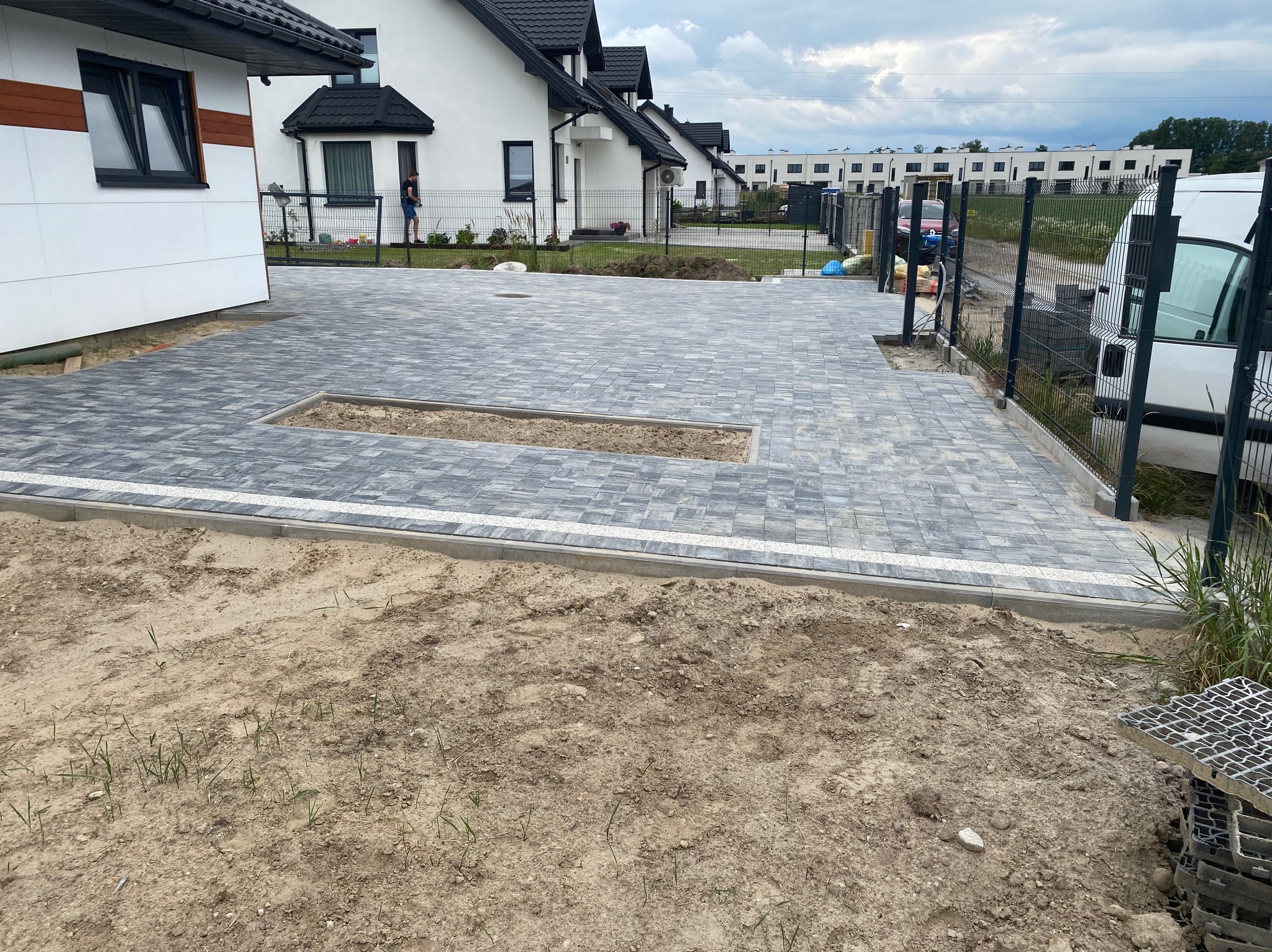 Nowo ułożona, szara kostka brukowa na podjeździe przed domem jednorodzinnym z przygotowanym miejscem na rabatę. Widoczne obrzeża i niezabudowany teren wokół.