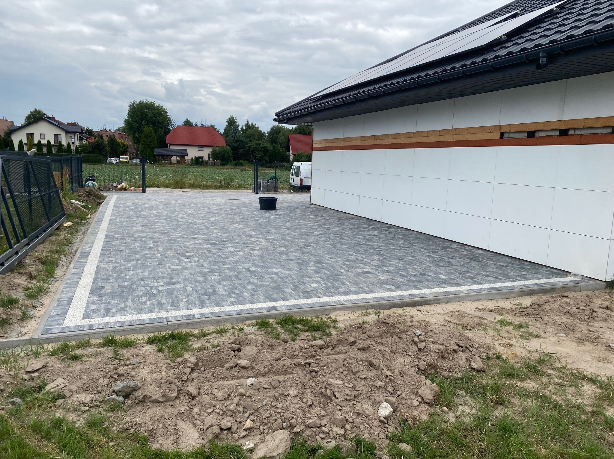 Ułożona szara kostka brukowa z jasnym obramowaniem na podjeździe przed nowoczesnym domem z panelami słonecznymi na dachu, w tle widoczne inne domy i pochmurne niebo.