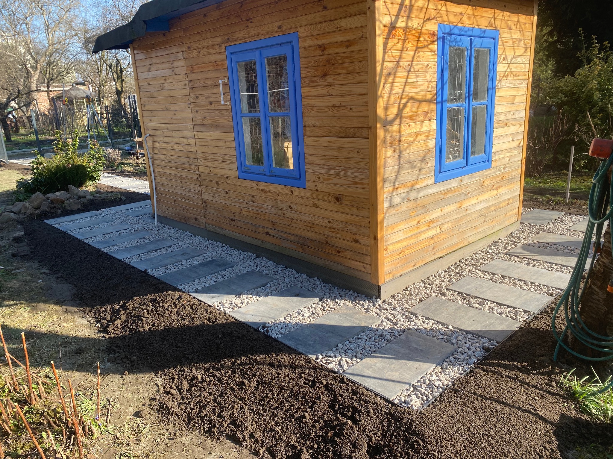 Ścieżka z szarych płyt ułożona wokół drewnianego domku ogrodowego, wysypana białym grysem, z widoczną świeżo przekopaną ziemią po bokach.