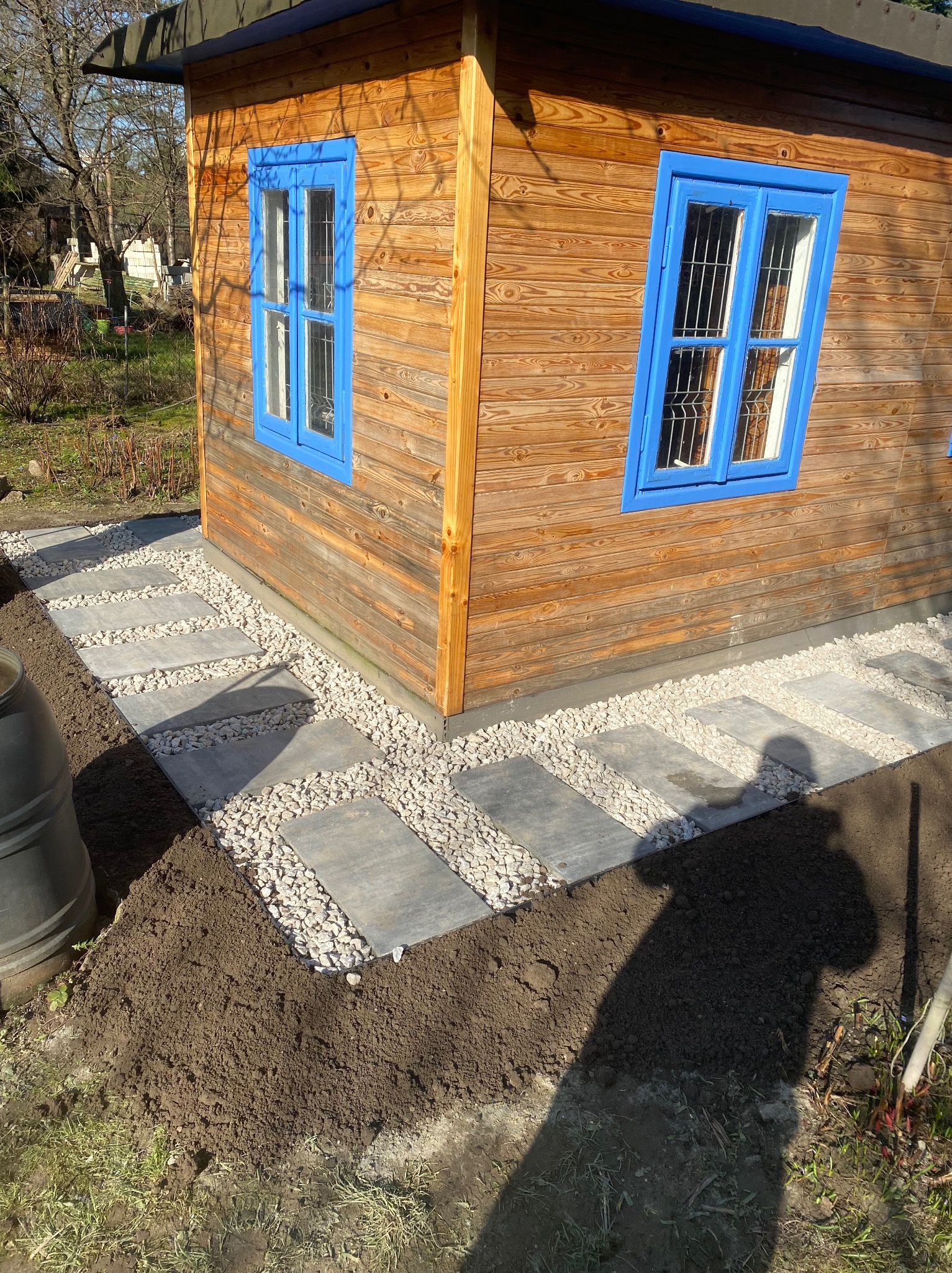 Ścieżka z szarych płyt chodnikowych ułożona wokół drewnianego domku z niebieskimi oknami, wypełniona białym grysem, obok świeżo przekopana ziemia.