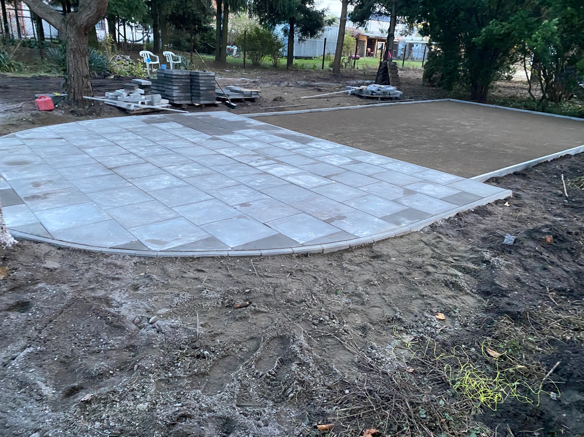 Częściowo ułożona, szara kostka brukowa w kształcie półkola, sąsiadująca z przygotowanym podłożem z piasku, w tle drzewa i ogrodzenie.