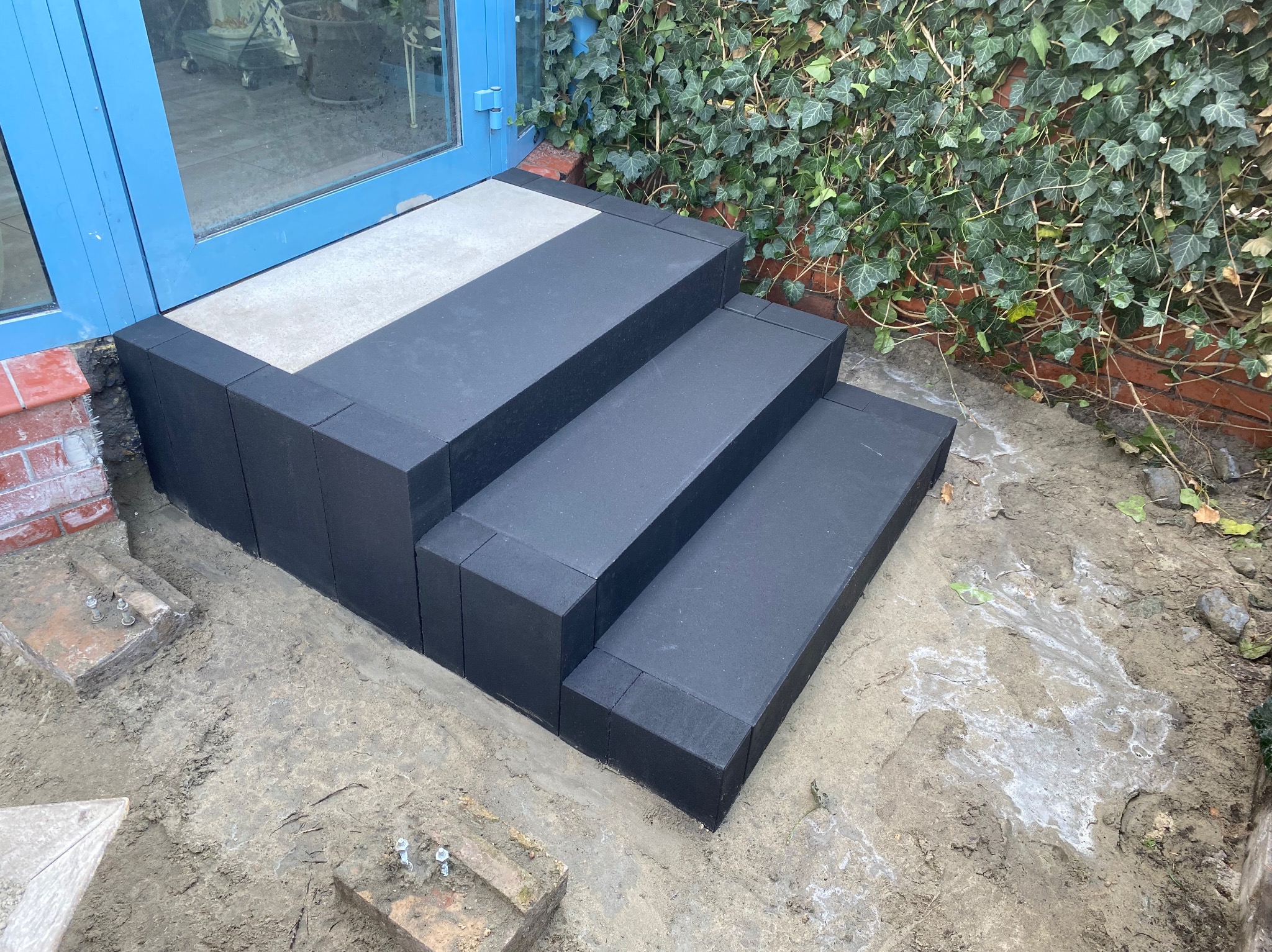 Trzy stopnie z ciemnej kostki prowadzące do niebieskich drzwi, z widocznymi śladami montażu i roślinnością w tle.