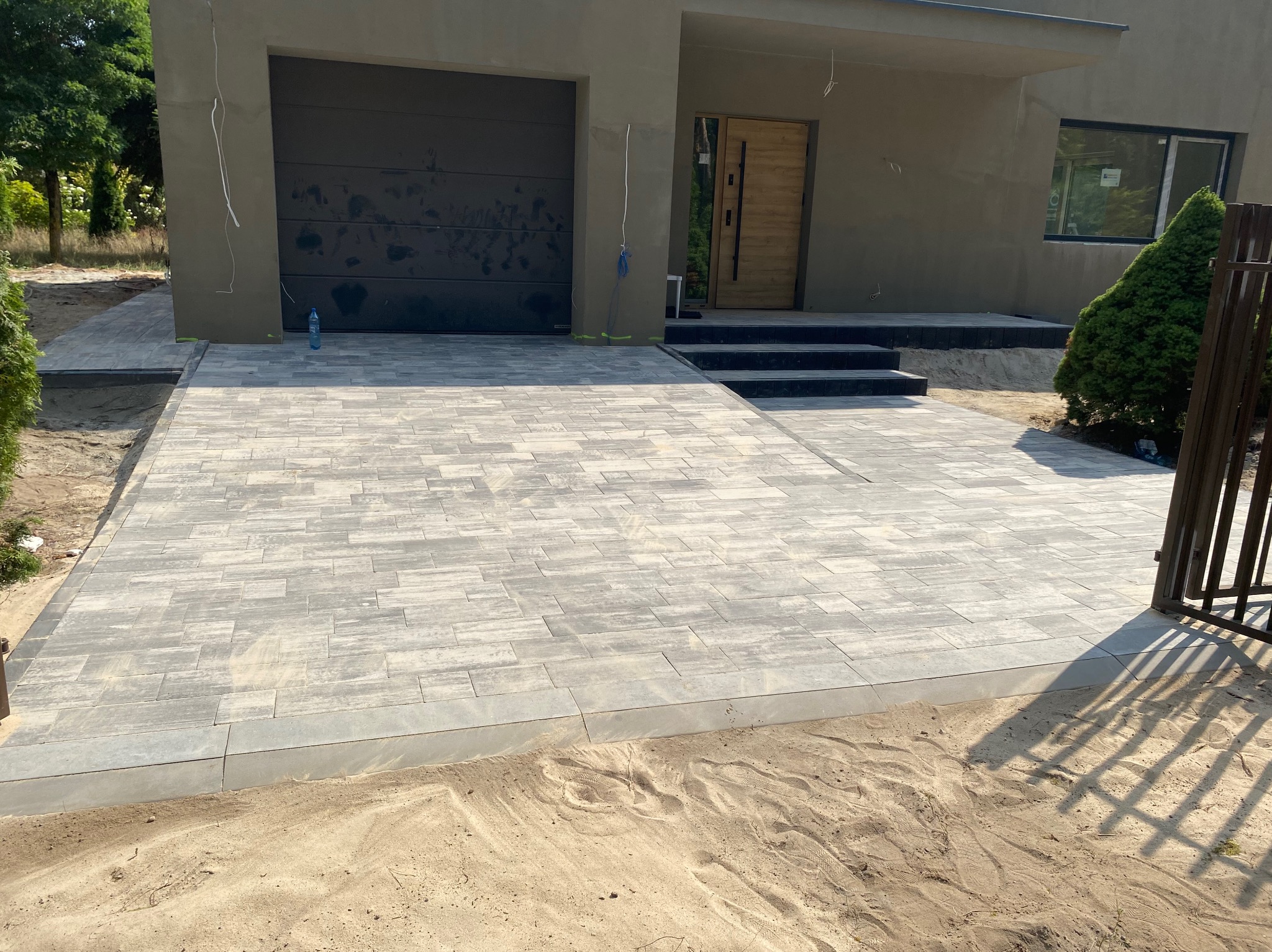 Nowo ułożony, szary chodnik z kostki brukowej przed nowoczesnym domem z garażem i drewnianymi drzwiami wejściowymi, widoczne obrzeża i piasek.