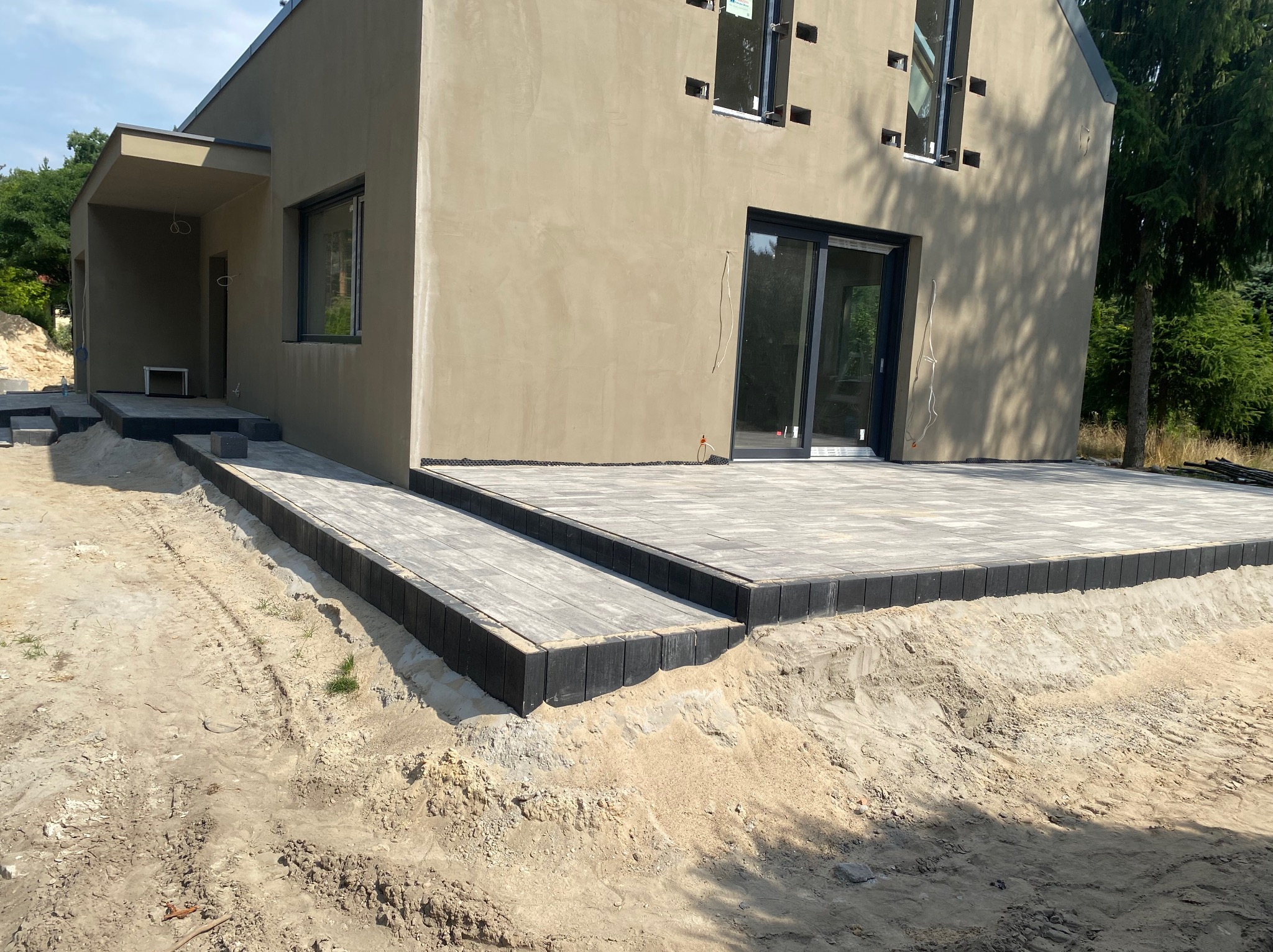 Nowoczesny dom z tarasem z jasnoszarej kostki brukowej z czarnym obrzeżem, teren wokół w trakcie prac ziemnych.