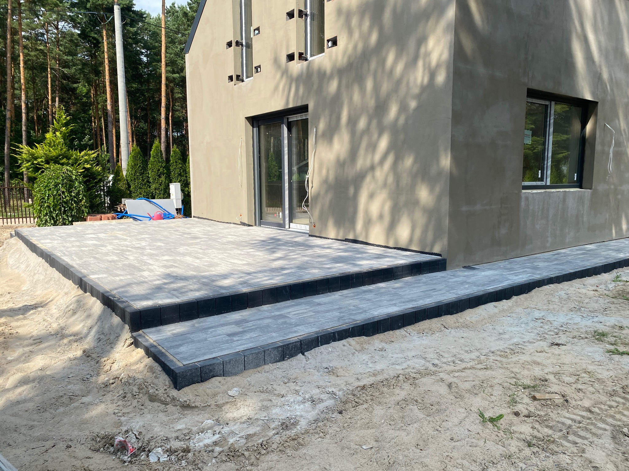 Nowo ułożony taras z szarej kostki brukowej z czarnym obrzeżem w Warszawie, prowadzący do budynku z dużym oknem i drzwiami balkonowymi, widoczne ślady prac ziemnych.