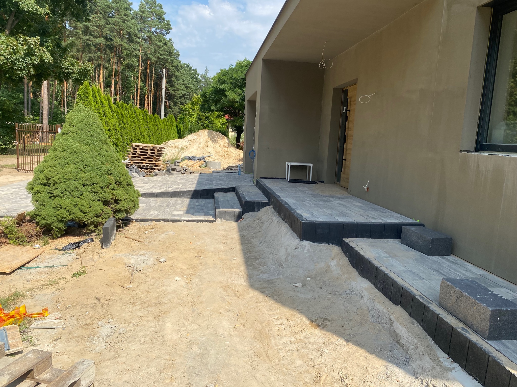 Fragment układania szarej kostki brukowej przed domem, widoczne schody i przygotowany teren, w tle drzewa i sterta piasku.