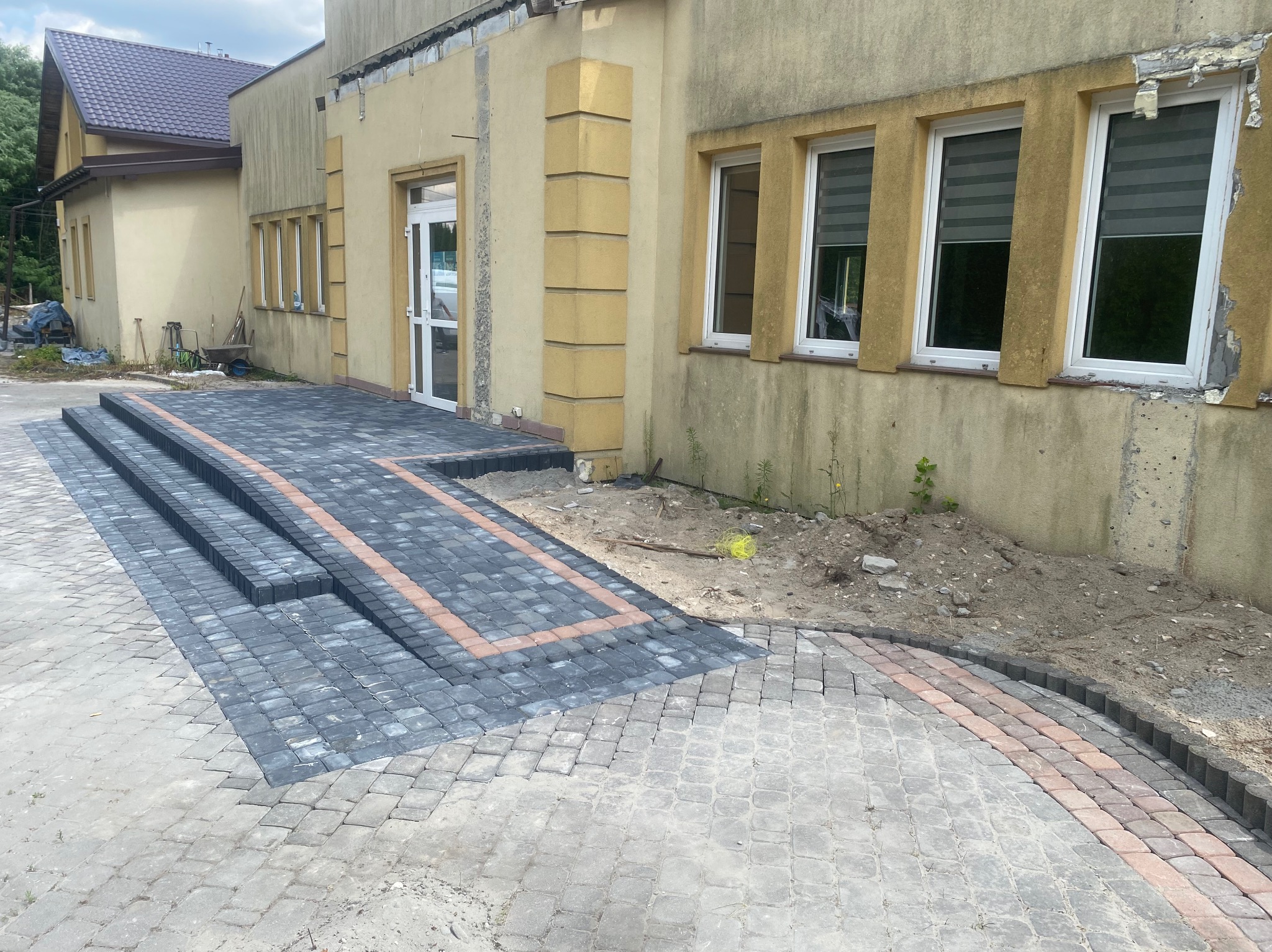 Ułożona szara kostka brukowa z obramowaniem z czerwonej kostki prowadząca do budynku z beżową elewacją i białymi oknami, częściowo widoczne schody z kostki.