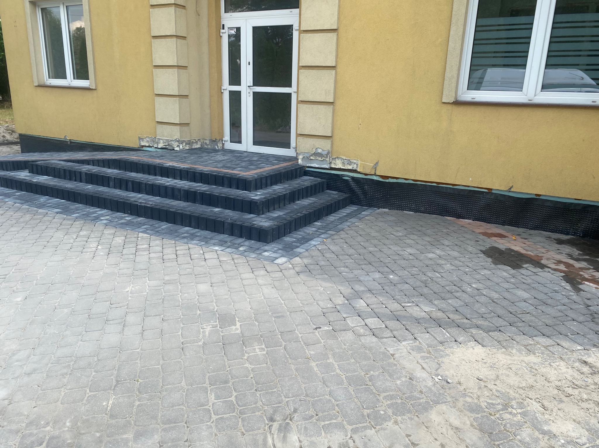 Ułożona szara kostka brukowa przed budynkiem z żółtą elewacją i białymi oknami, widoczne trzy stopnie schodów z ciemnoszarej kostki, częściowo widoczna folia izolacyjna przy ścianie.