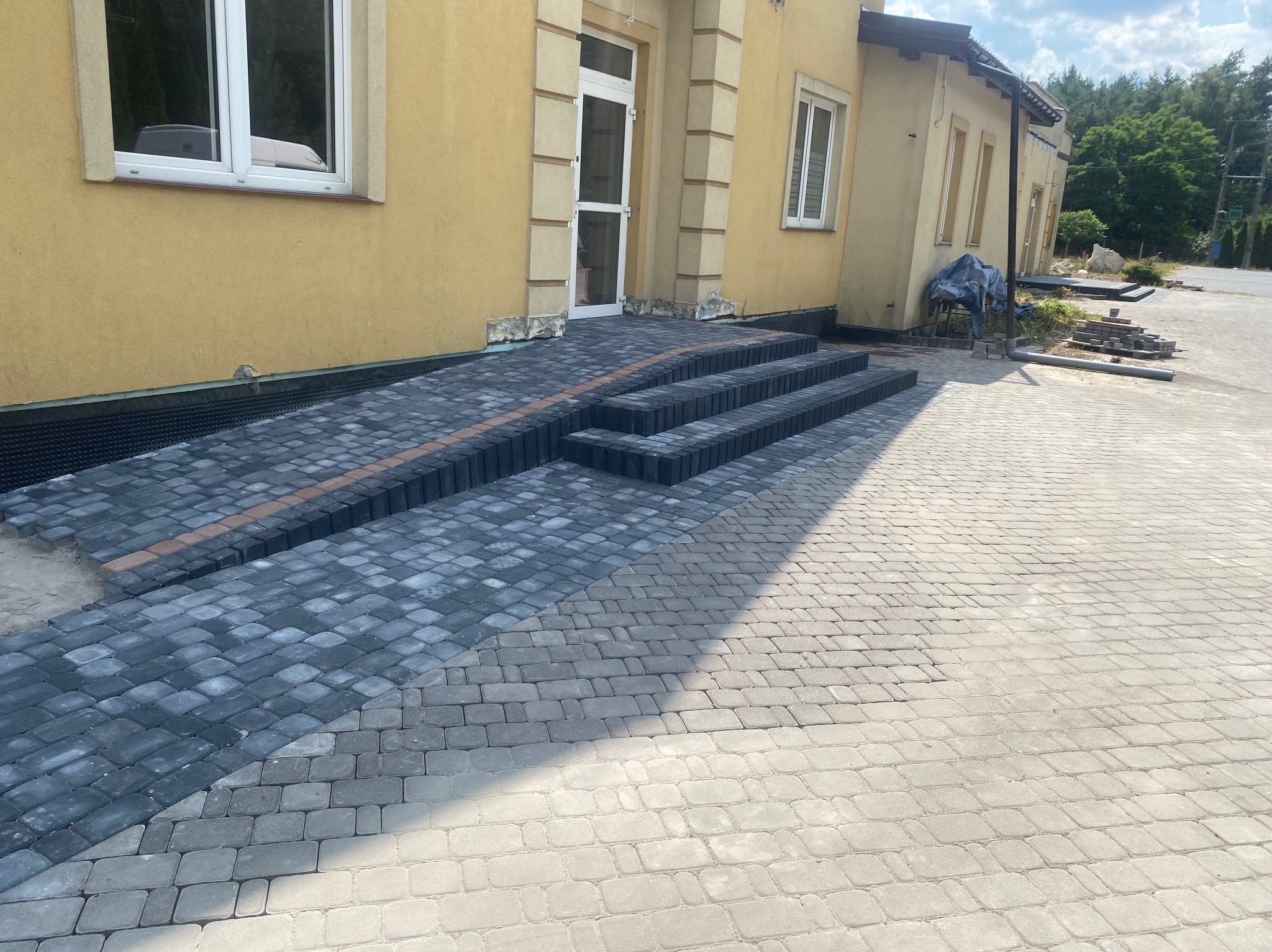 Widok na schody i podjazd z kostki brukowej w kolorze szarym i czarnym, prowadzące do budynku z żółtą elewacją, częściowo ukończone.