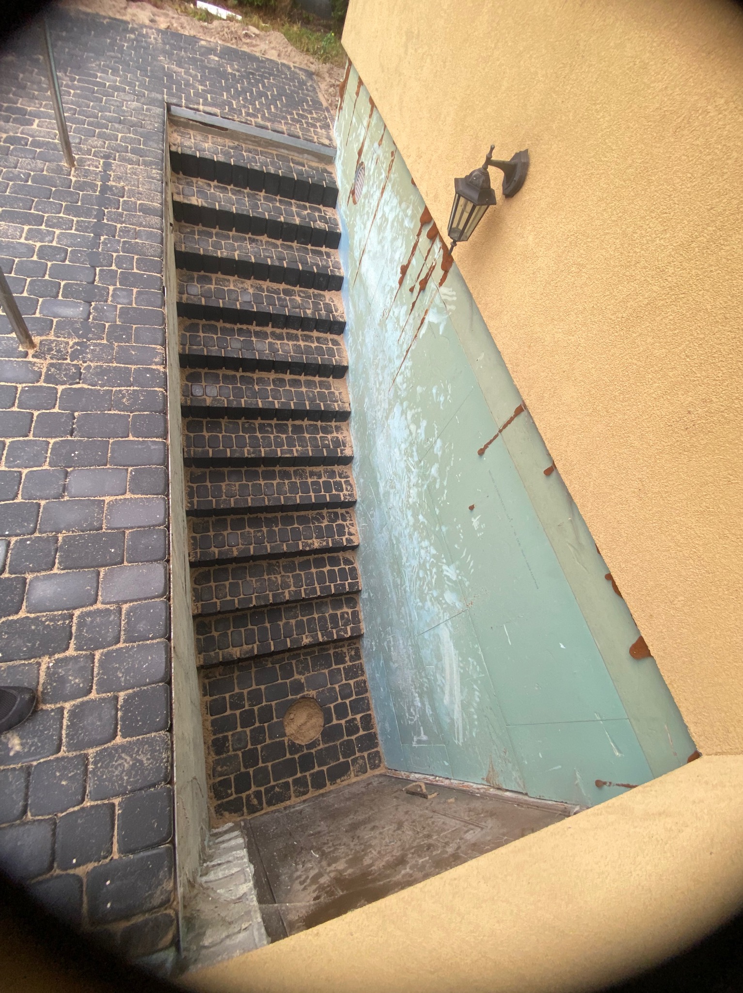 Schody z ciemnej kostki brukowej prowadzące w dół, widok z góry, obok ściana budynku z lampą.