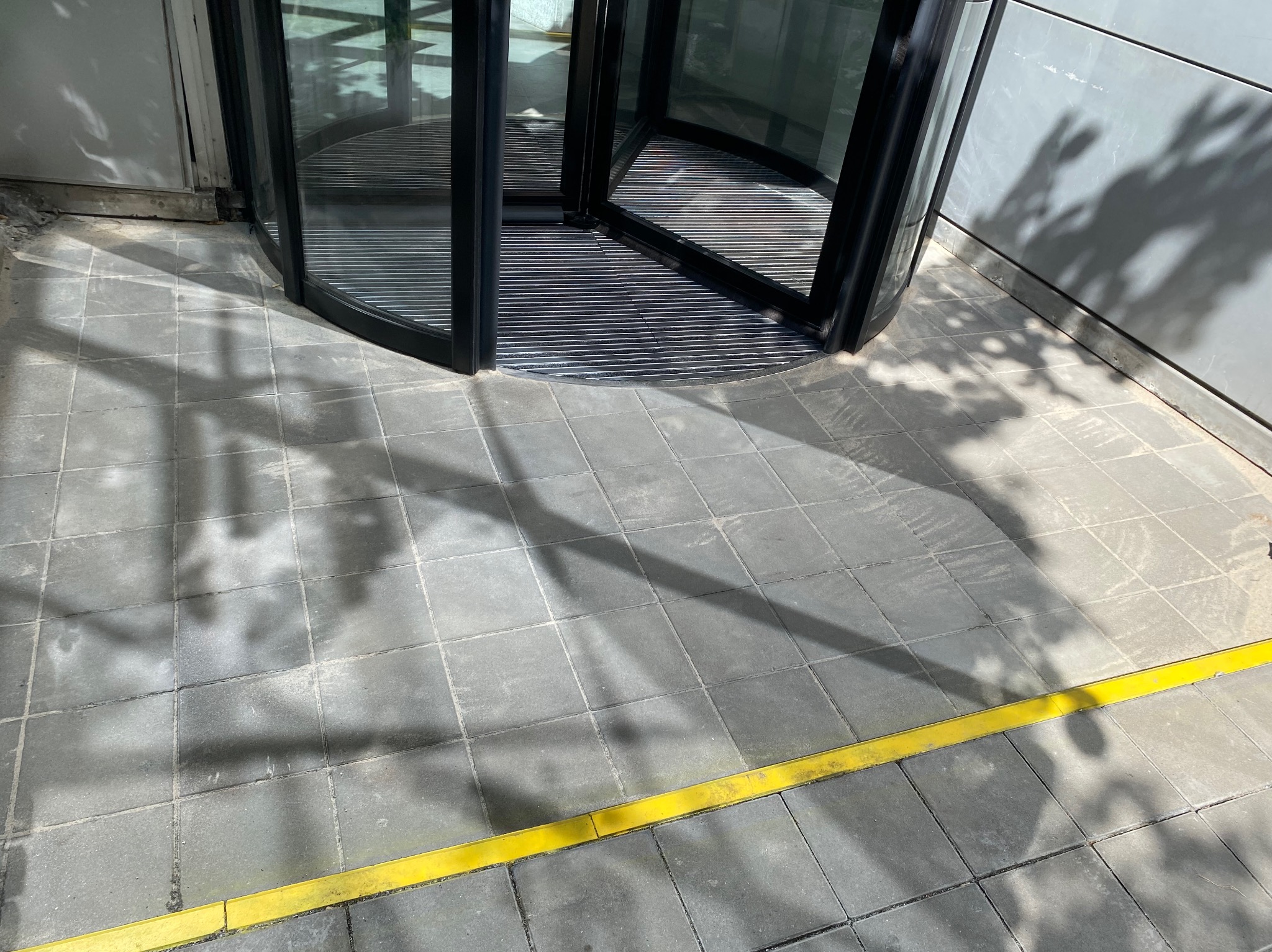 Szare płytki chodnikowe przy obrotowych drzwiach wejściowych, z widocznym żółtym obrzeżem i cieniami drzew.