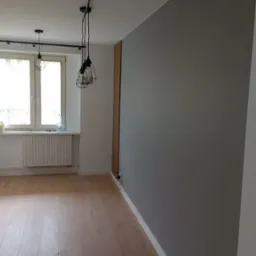 Szara ściana po malowaniu w nowocześnie urządzonym pokoju z panelami podłogowymi, widoczne lampy wiszące i dekoracyjna drewniana listwa na ścianie.