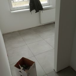 Daniel wypych - Świeżo ułożona, jasnoszara podłoga z płytek, z krzyżykami dystansowymi w spoinach, karton z narzędziami w rogu pokoju, kurtka wisząca na oknie w tle.
