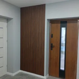 Wnętrze przedpokoju z drewnianą ścianką lamelową, brązowymi drzwiami wejściowymi z szybą i białymi drzwiami wewnętrznymi, szare ściany.