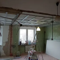 Wnętrze w trakcie montażu sufitu podwieszanego z widoczną konstrukcją metalową, workami z zaprawą na drabinie i odsłoniętymi fragmentami ścian z zieloną farbą.