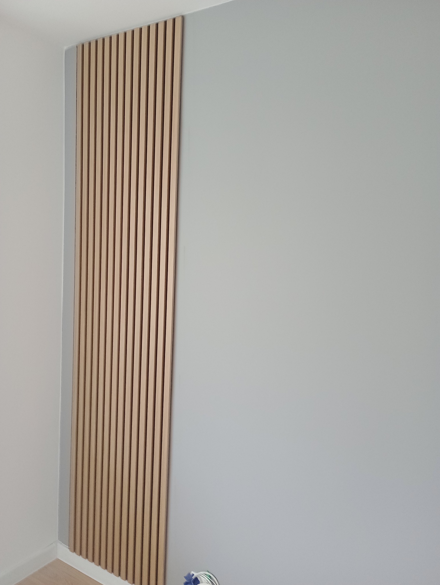 Fragment ściany z zamontowanymi pionowymi panelami dekoracyjnymi z jasnego drewna, przylegający do szarej ściany w rogu pomieszczenia.