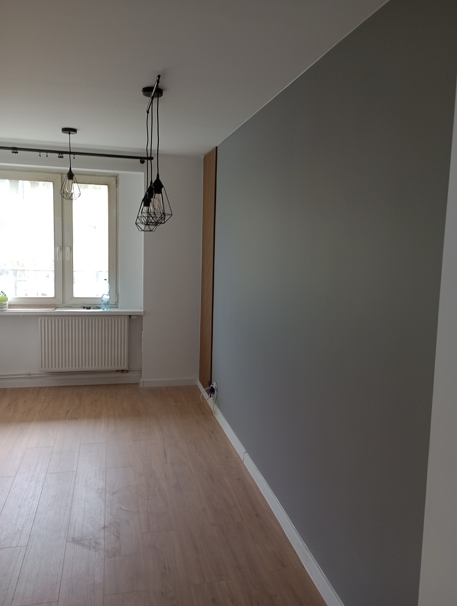 Szara ściana po malowaniu w nowocześnie urządzonym pokoju z panelami podłogowymi, widoczne lampy wiszące i dekoracyjna drewniana listwa na ścianie.