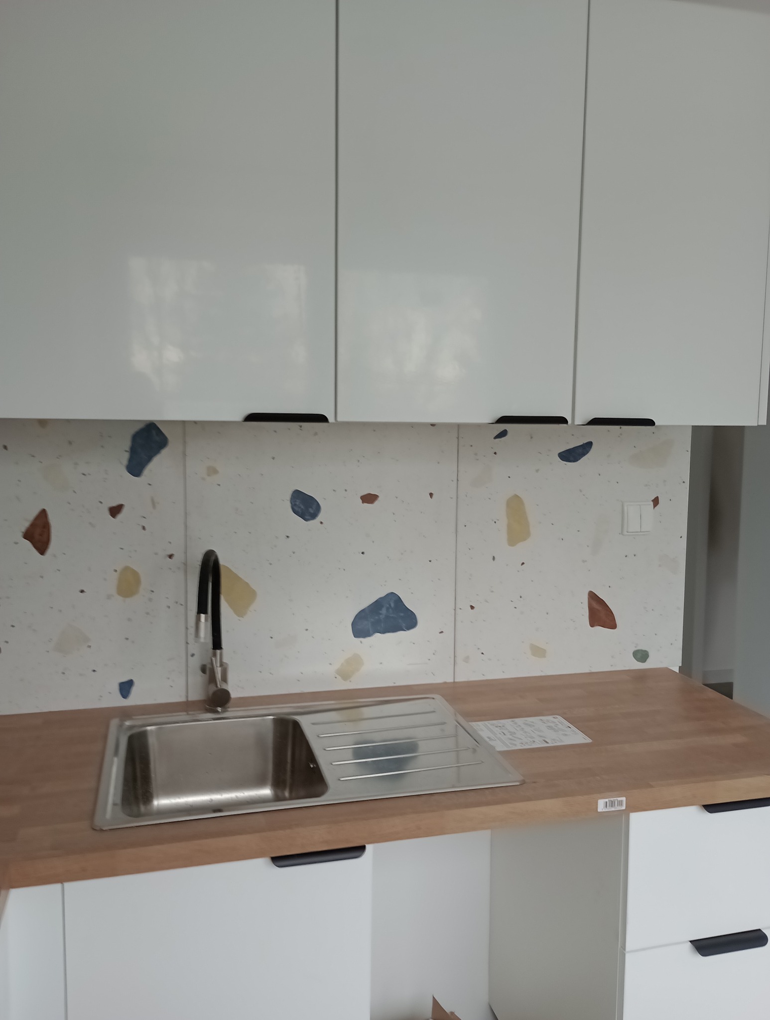 Nowoczesna kuchnia z białymi szafkami, drewnianym blatem i oryginalną ścianą nad blatem w stylu lastryko z kolorowymi plamkami. Widoczny zlewozmywak ze stali nierdzewnej i czarna bateria.