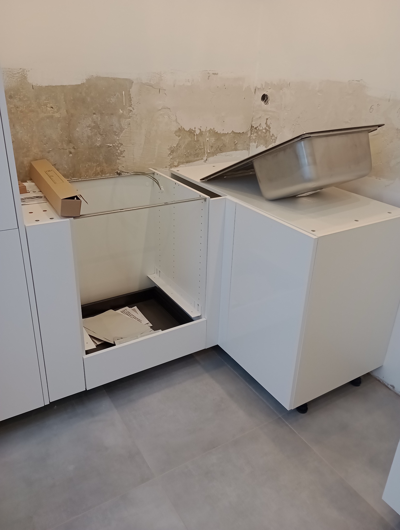 Montaż białych szafek kuchennych w surowym stanie, widoczna ściana przed gładzią i zlew tymczasowo umieszczony na blacie.