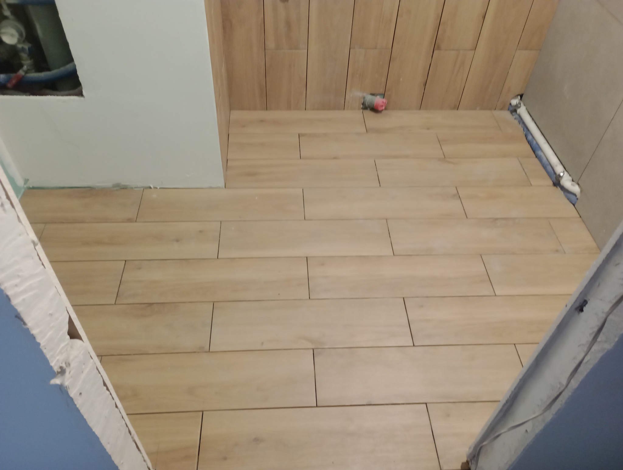 Układanie podłogowych płytek imitujących drewno w łazience, widoczne fragmenty ścian i instalacji hydraulicznej w trakcie remontu.