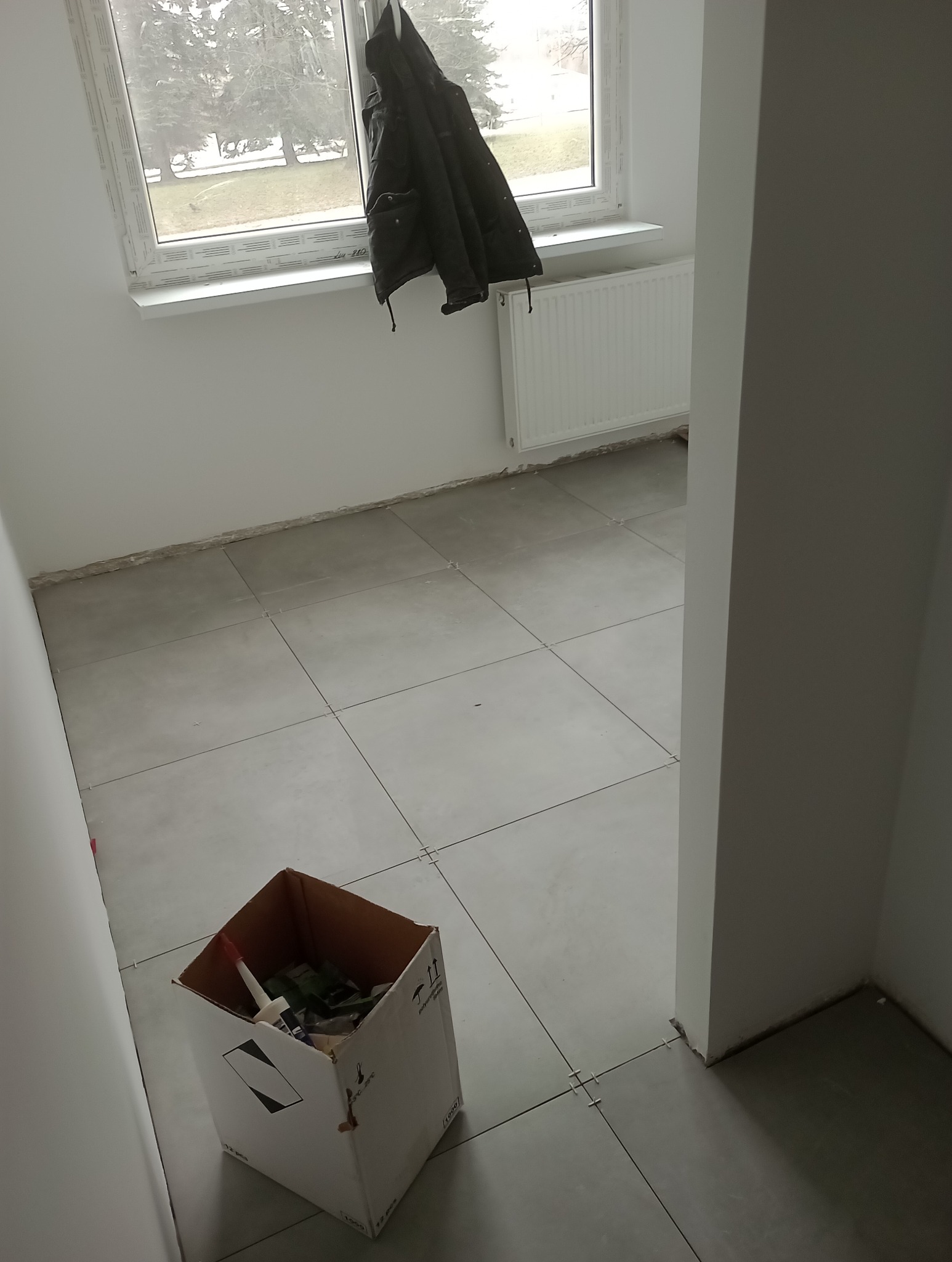 Świeżo ułożona, jasnoszara podłoga z płytek, z krzyżykami dystansowymi w spoinach, karton z narzędziami w rogu pokoju, kurtka wisząca na oknie w tle.