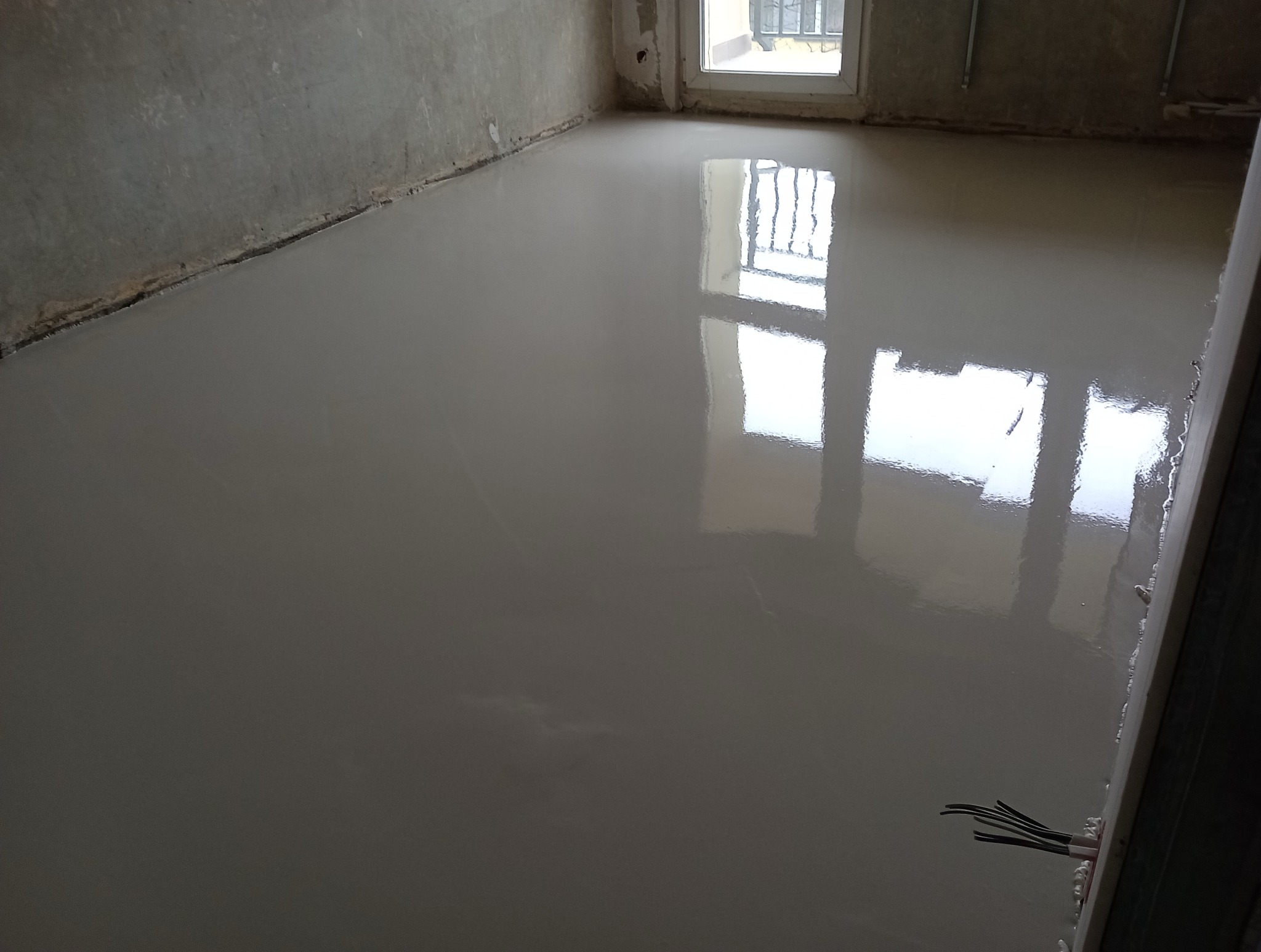 Świeżo wylana, samopoziomująca wylewka podłogowa w pomieszczeniu w Jodłowej, z widocznym odbiciem okna w gładkiej powierzchni.