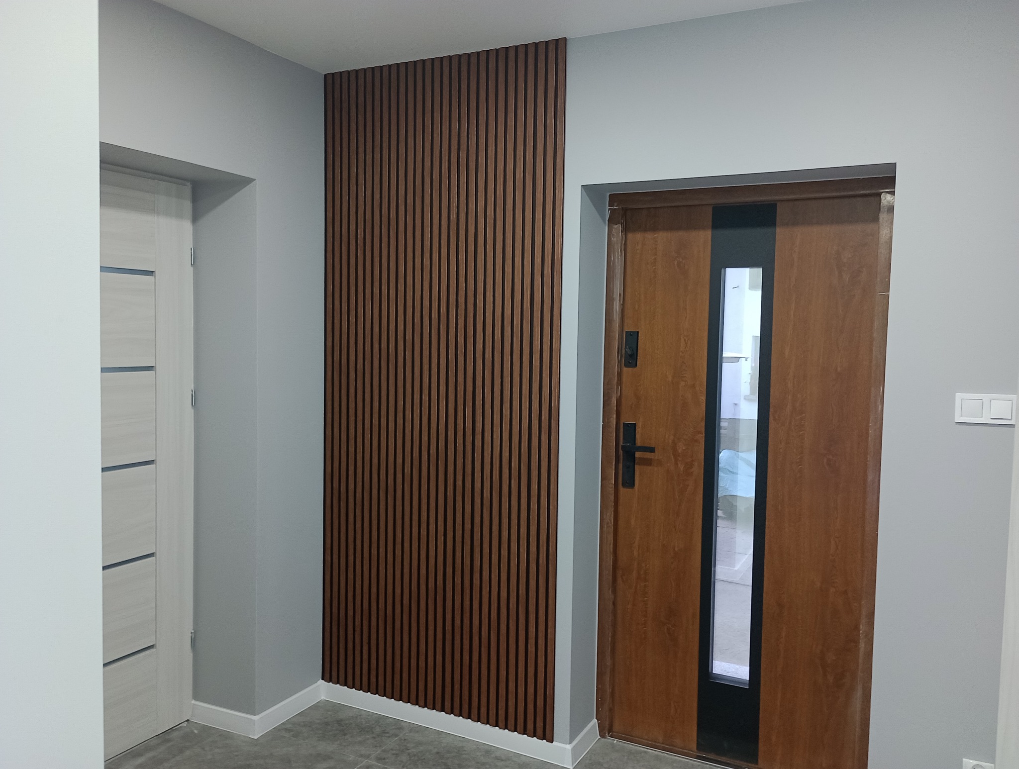 Wnętrze przedpokoju z drewnianą ścianką lamelową, brązowymi drzwiami wejściowymi z szybą i białymi drzwiami wewnętrznymi, szare ściany.