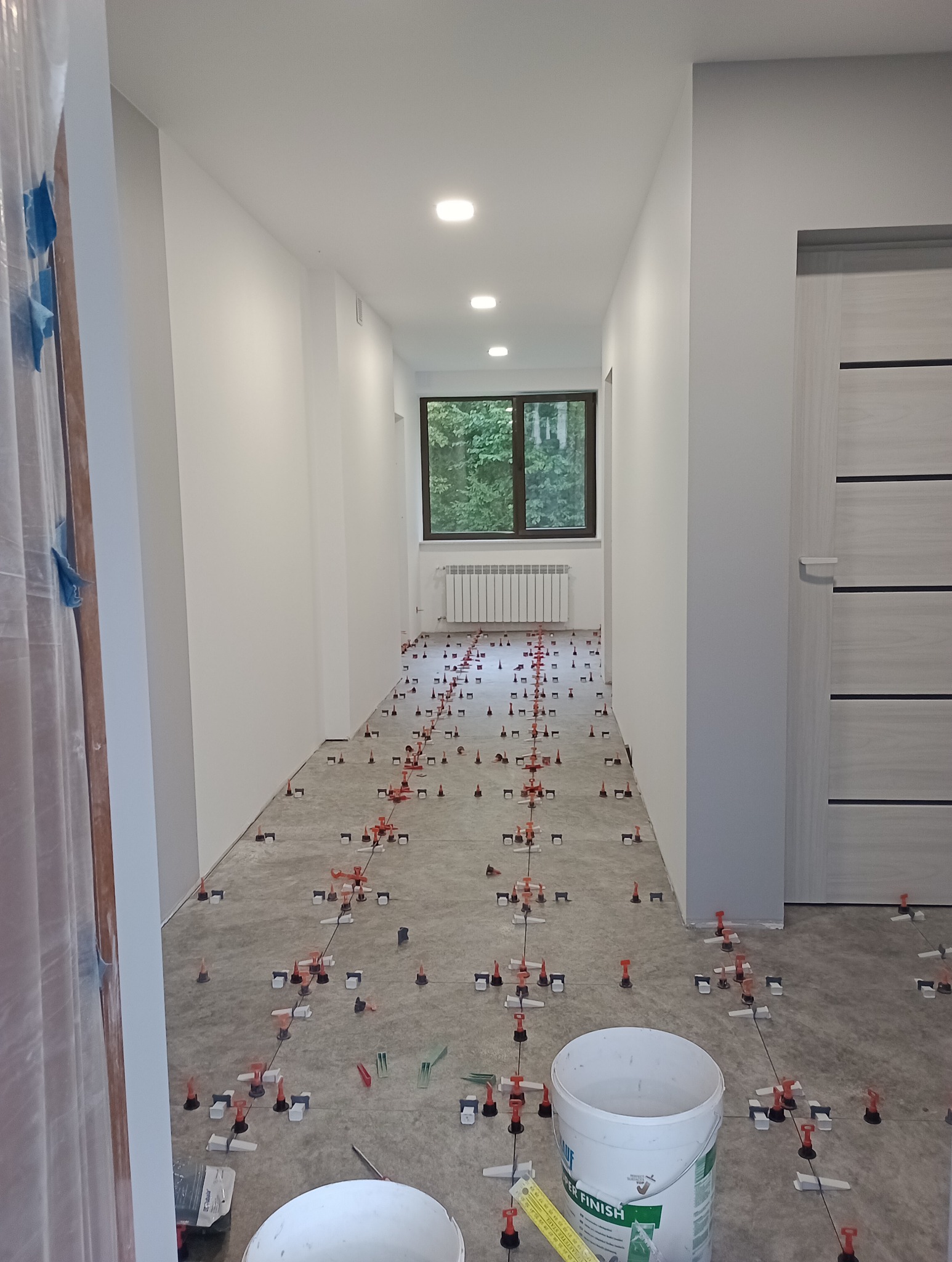 Korytarz w trakcie układania płytek podłogowych z użyciem systemu poziomowania; widoczne białe ściany, szare drzwi z poziomymi paskami, okno z widokiem na drzewa, grzejnik oraz dwa białe wiadra...