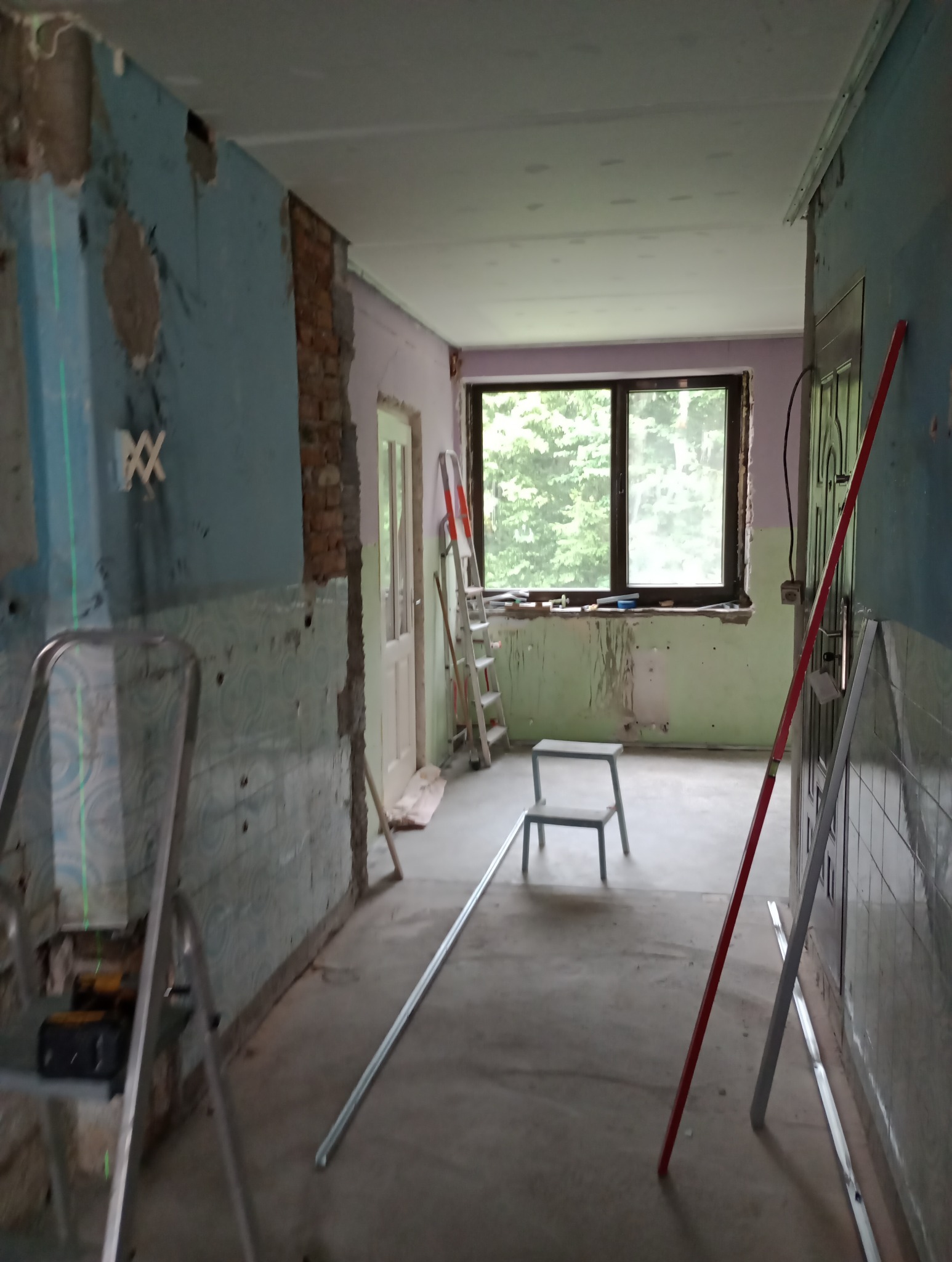 Wnętrze w trakcie remontu: odsłonięta cegła, resztki starej farby na ścianach, drabiny, poziomica laserowa i profile do płyt karton-gips, okno z widokiem na zieleń.