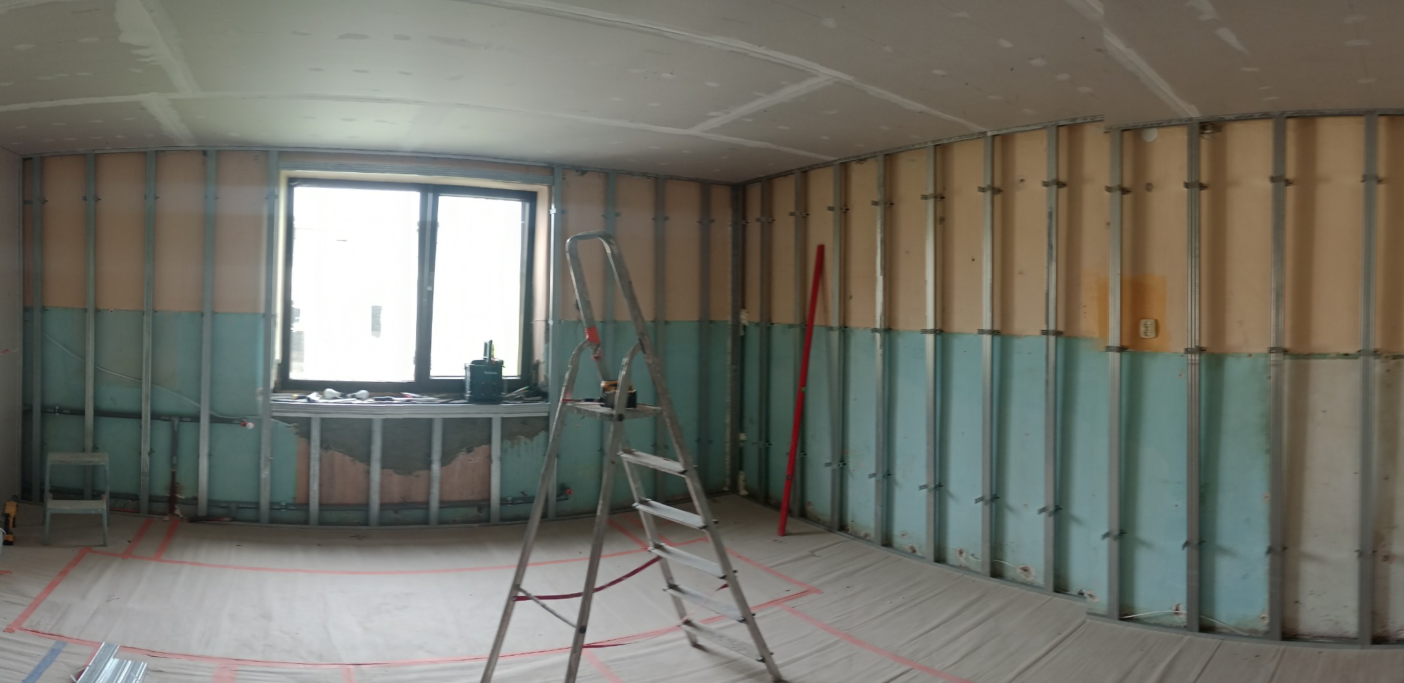 Wnętrze w trakcie prac z płytami kartonowo-gipsowymi: widoczne metalowe profile, częściowo zamontowane płyty, zakryta podłoga, drabina i okno z narzędziami.