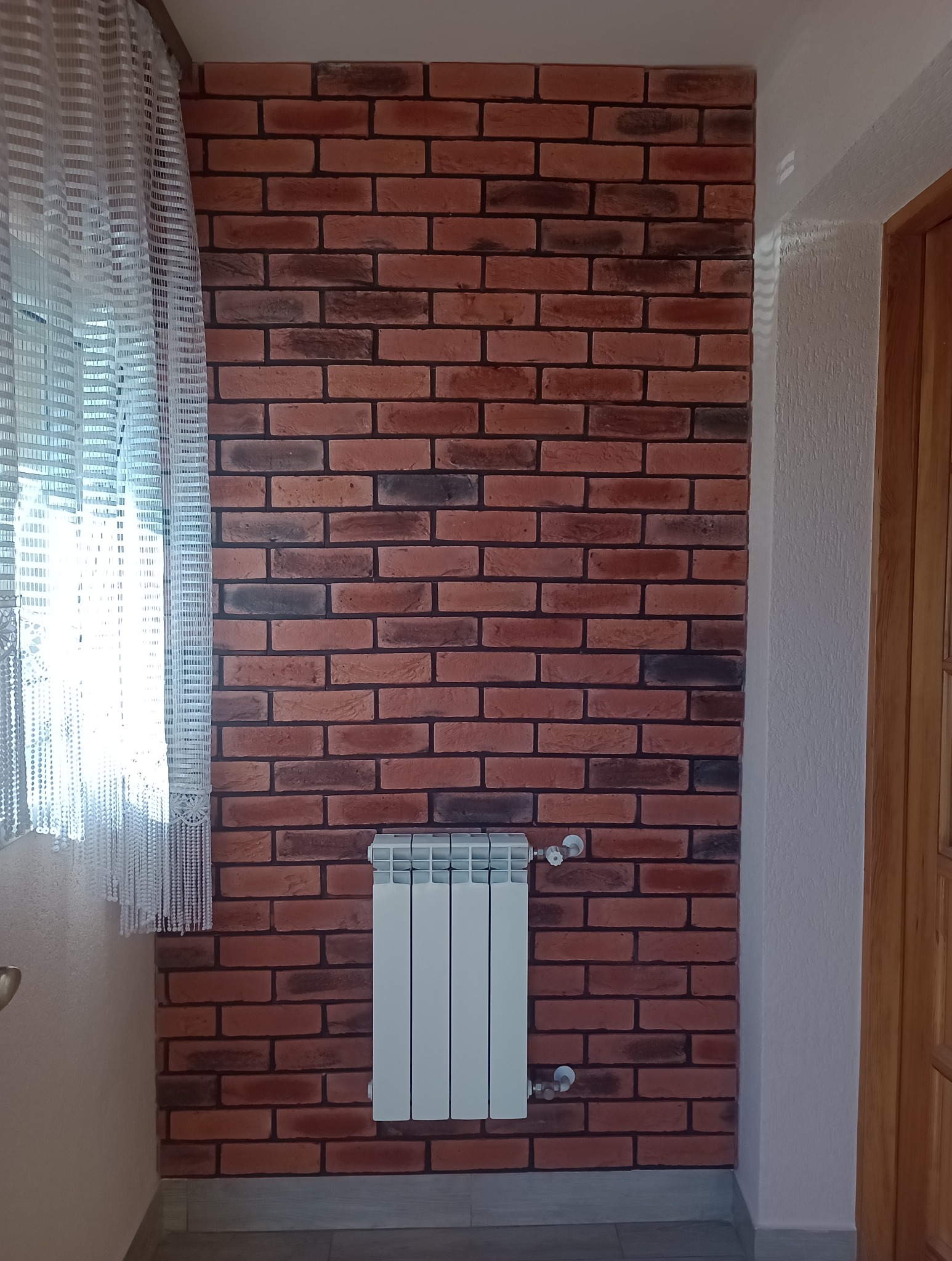 Ściana wyłożona cegłą dekoracyjną w odcieniach czerwieni i brązu, z zamontowanym białym grzejnikiem aluminiowym. Obok widoczna biała firanka i fragment drewnianych drzwi.