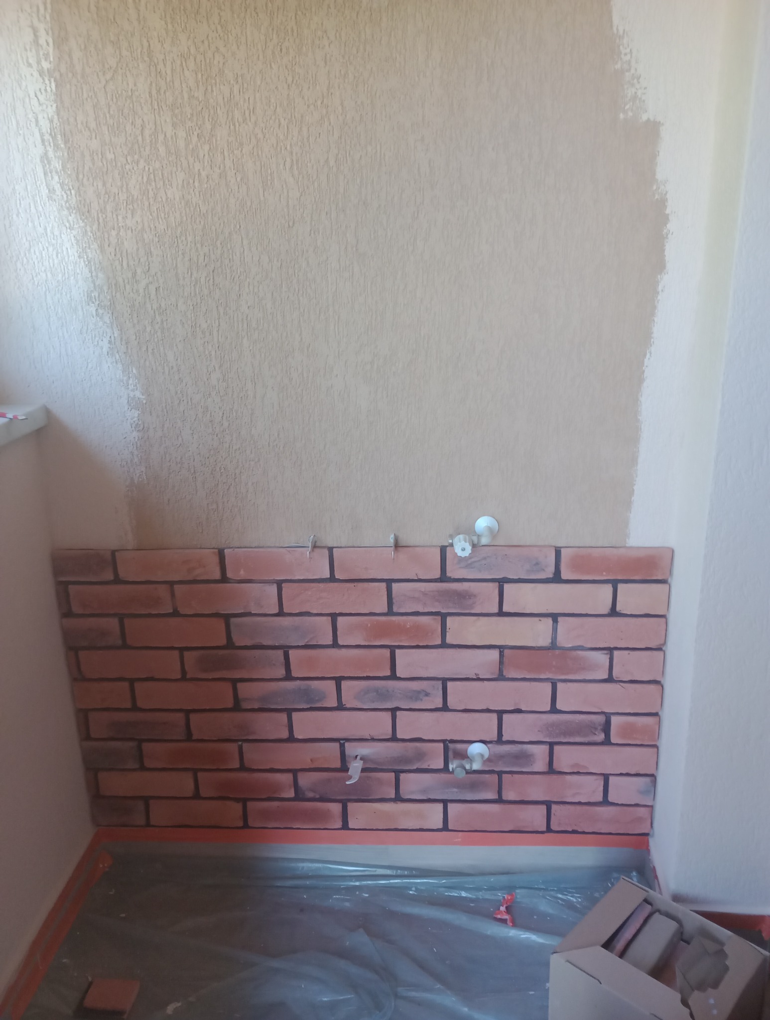 Fragment ściany z imitacją cegły w trakcie montażu, widoczne elementy mocujące, surowa ściana powyżej z śladami malowania, zabezpieczona folią podłoga.