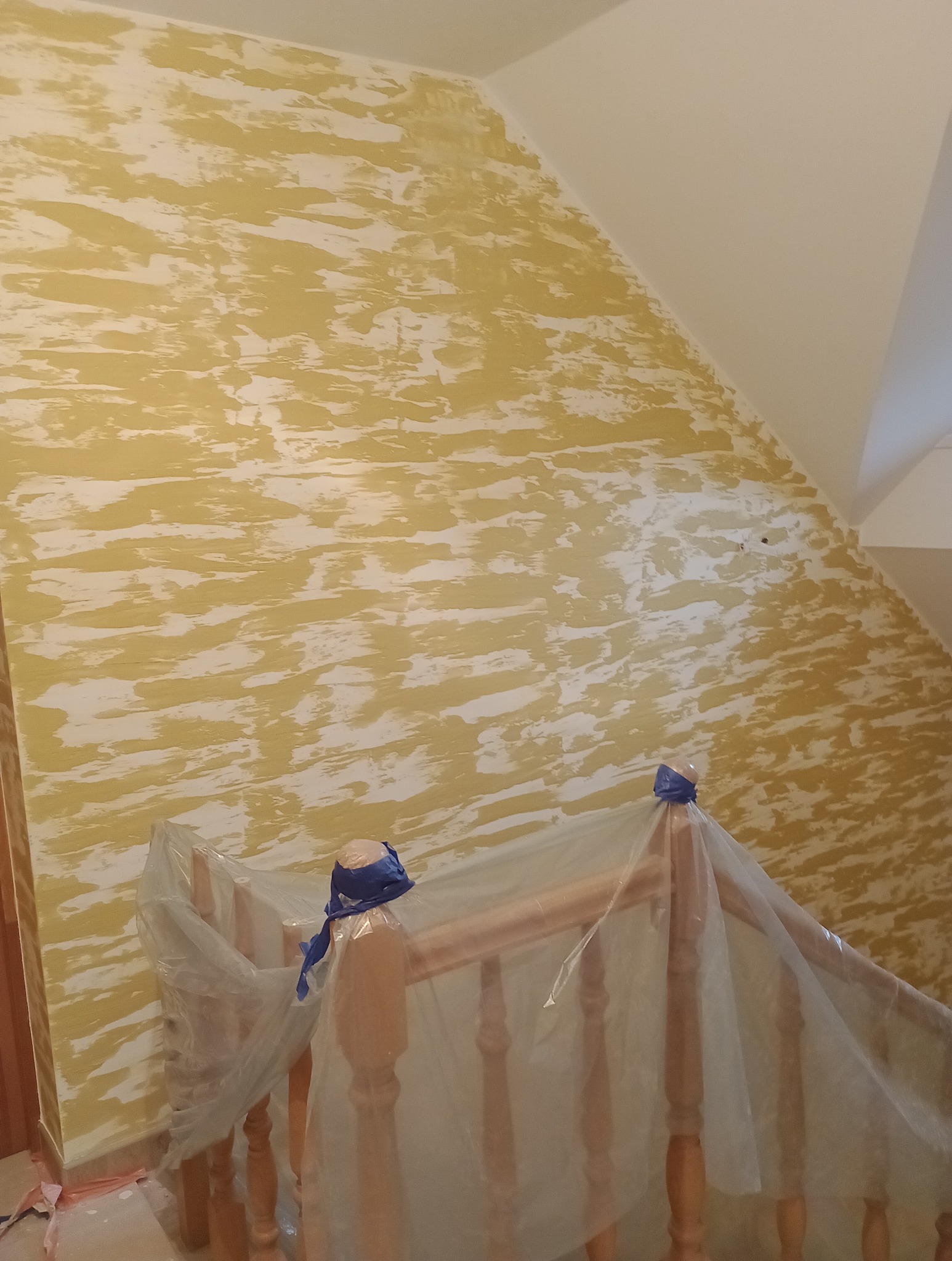 Ściana w trakcie malowania dekoracyjnego z efektem smug w odcieniach żółci i bieli, zabezpieczona folią drewniana balustrada schodów.