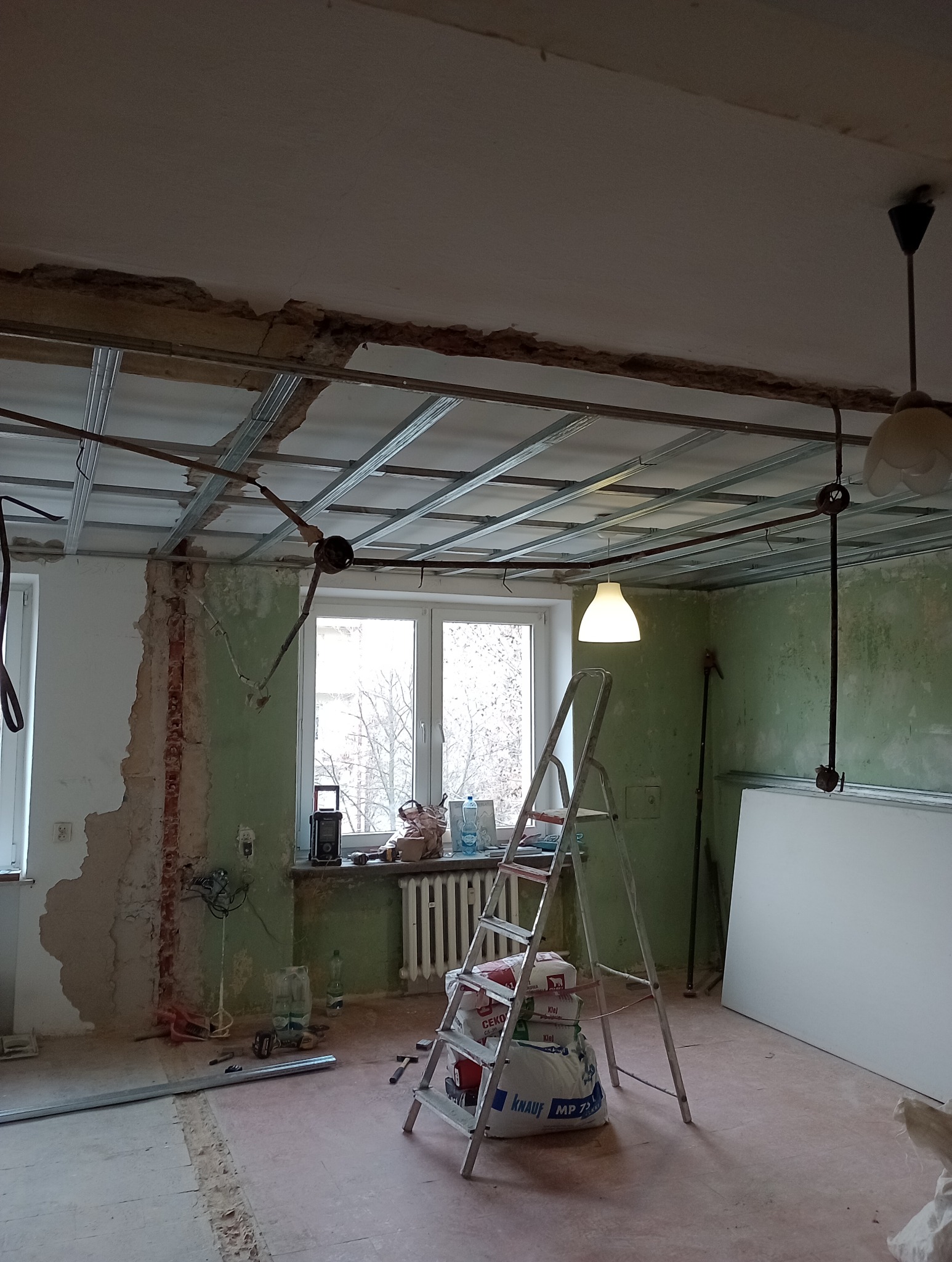 Wnętrze w trakcie montażu sufitu podwieszanego z widoczną konstrukcją metalową, workami z zaprawą na drabinie i odsłoniętymi fragmentami ścian z zieloną farbą.
