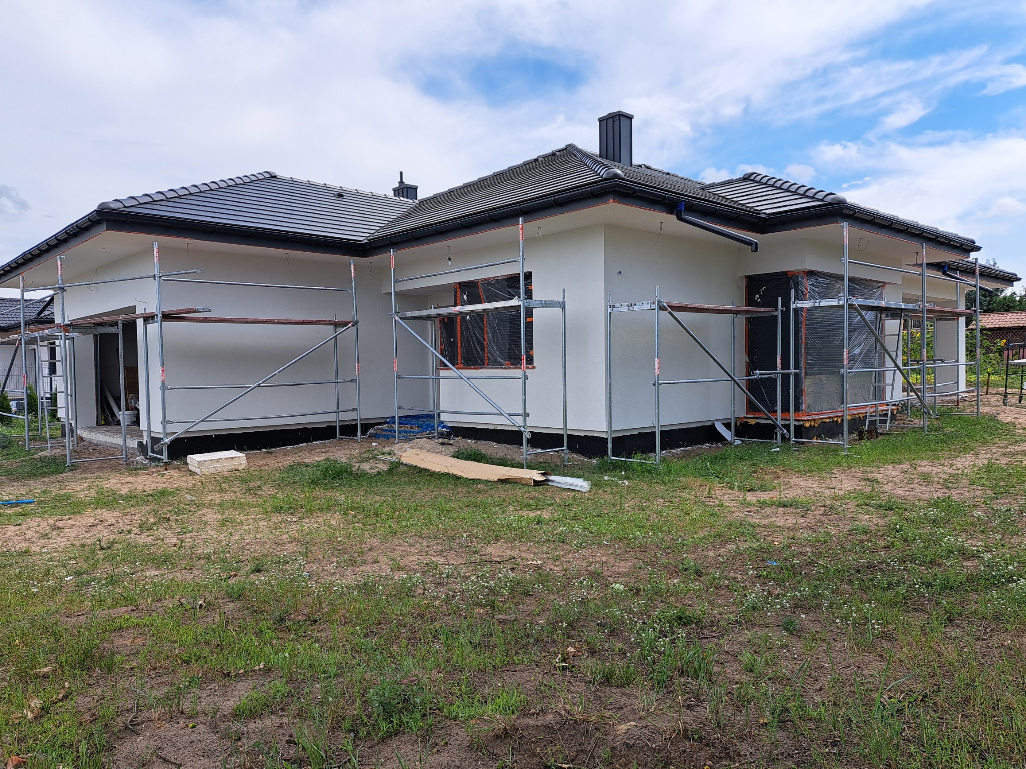 Dom jednorodzinny w trakcie ocieplania elewacji, widoczne rusztowania i zabezpieczone okna folią, czarna dachówka i komin, trawnik z młodą trawą.