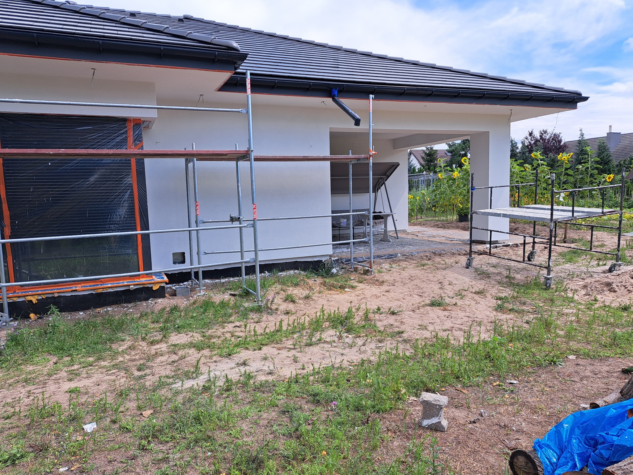 Dom jednorodzinny w Solcu Kujawskim w trakcie prac elewacyjnych, widoczne rusztowania, okno zabezpieczone folią, fragment ocieplenia, dachówka ceramiczna i słoneczniki w tle.