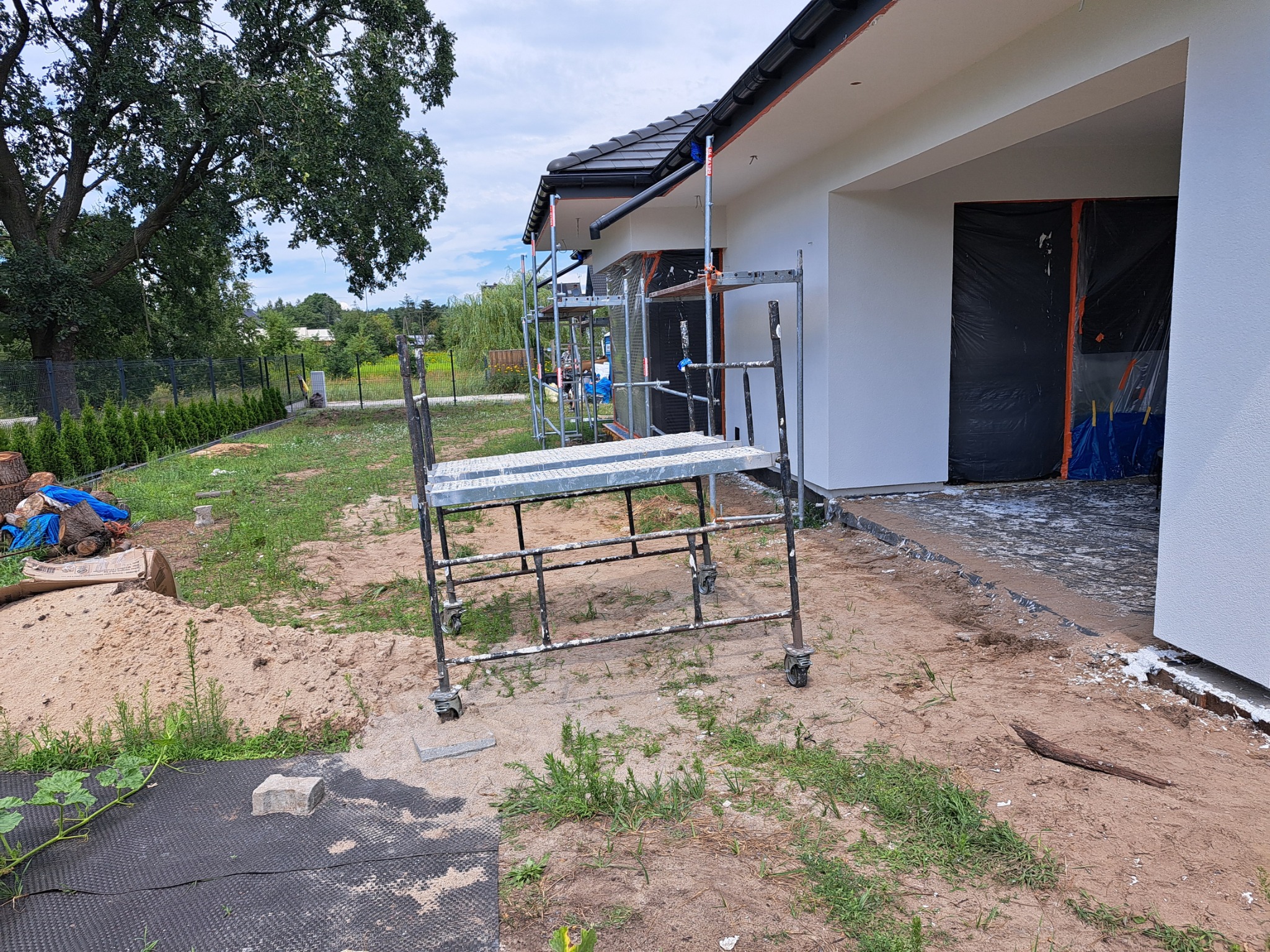 Dwukondygnacyjny dom jednorodzinny z białą elewacją w trakcie prac, widoczne rusztowania z resztkami farby i zabezpieczone okno folią, teren wokół w stanie surowym.