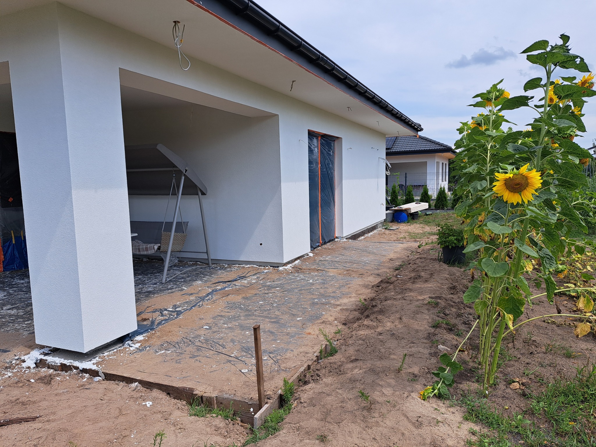 Nowoczesny dom jednorodzinny z białą elewacją, częściowo osłoniętym wejściem i świeżo otynkowanym filarem, z widocznymi śladami zaprawy na ziemi i osłoniętymi folią oknami, oraz wysokimi...