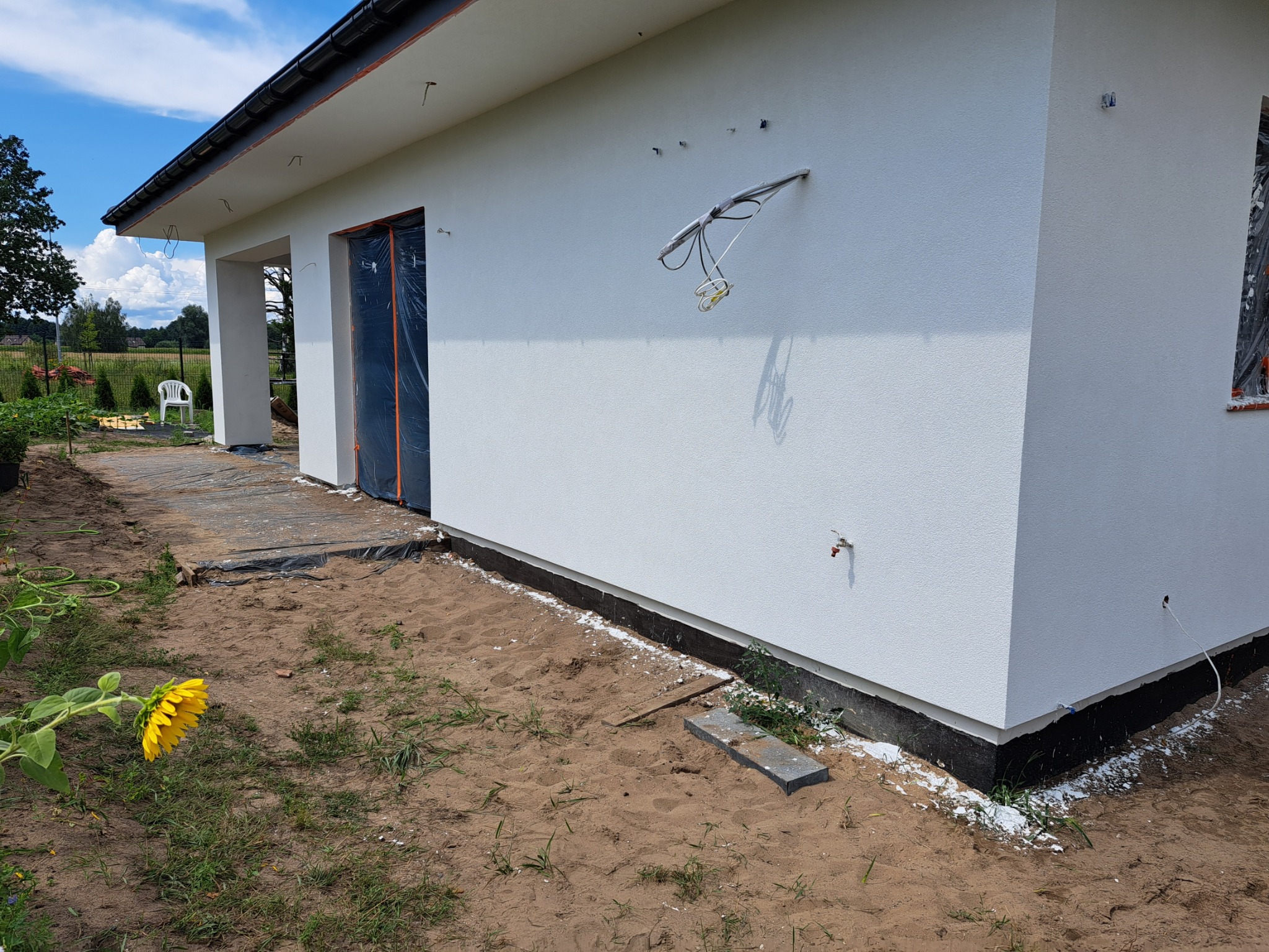 Świeżo otynkowany, biały dom jednorodzinny z widocznymi wystającymi kablami elektrycznymi i zabezpieczonymi folią otworami okiennymi i drzwiowymi, na piaszczystym terenie z kwitnącym słonecznikiem...