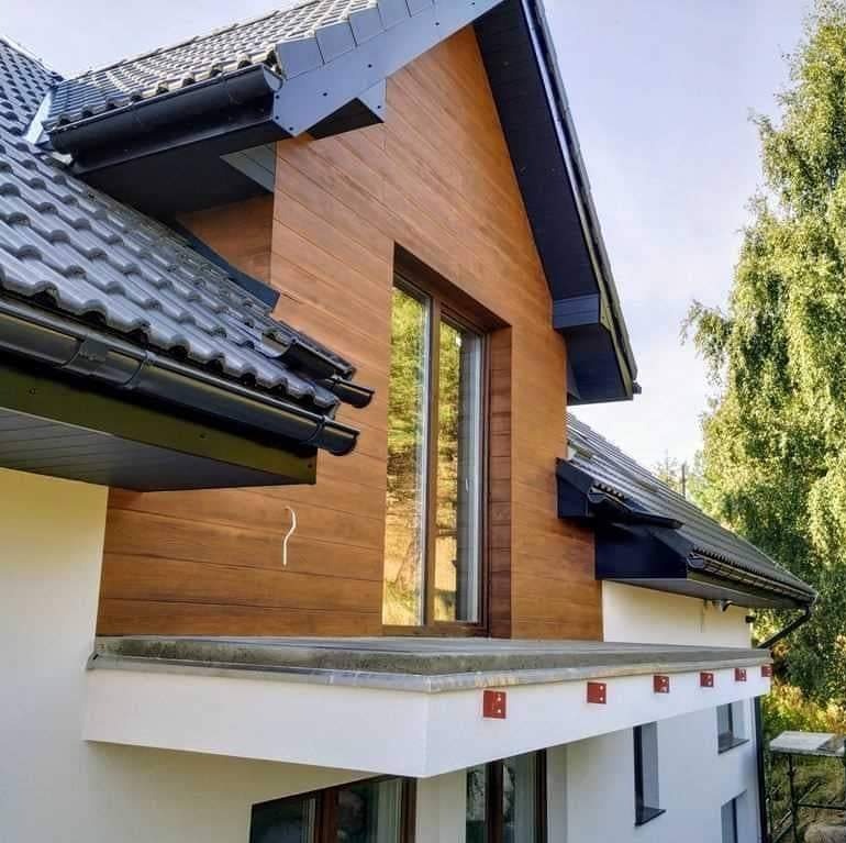Nowoczesny dom z elewacją z drewnopodobnych paneli, grafitowa dachówka i orynnowanie, widoczne czerwone elementy mocujące na balkonie.