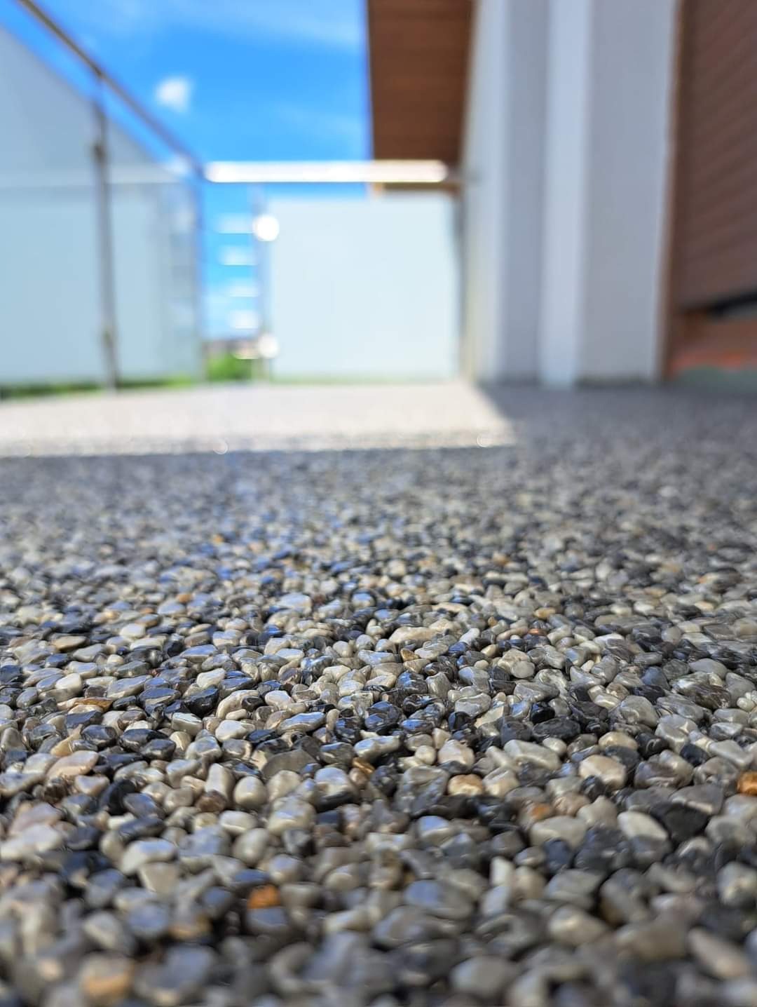 Posadzka z żywicy epoksydowej i drobnych kamyczków na tarasie, widok z niskiej perspektywy, w tle szklana balustrada i fragment elewacji.