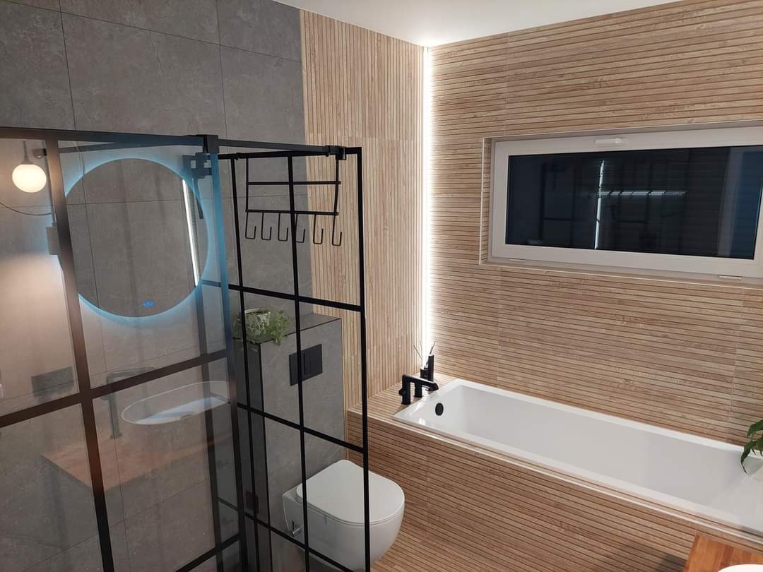 Nowoczesna łazienka z kabiną prysznicową typu walk-in z czarnymi profilami, okrągłym lustrem z podświetleniem LED, drewnopodobnymi płytkami ściennymi i białą wanną.