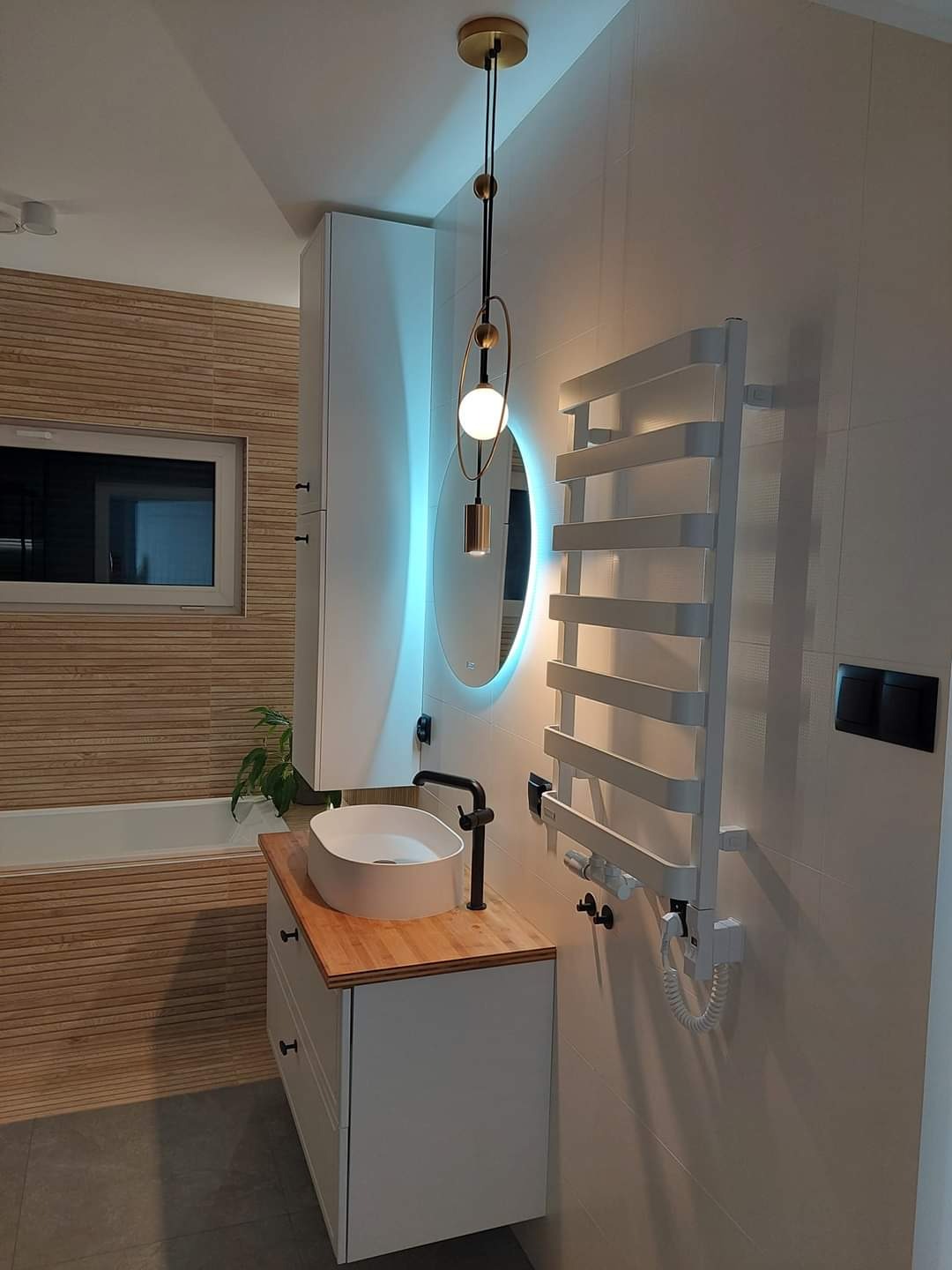 Nowoczesna łazienka z drewnianymi akcentami: owalna umywalka nablatowa na białej szafce z drewnianym blatem, podświetlane lustro, grzejnik drabinkowy i wisząca lampa z elementami złota.