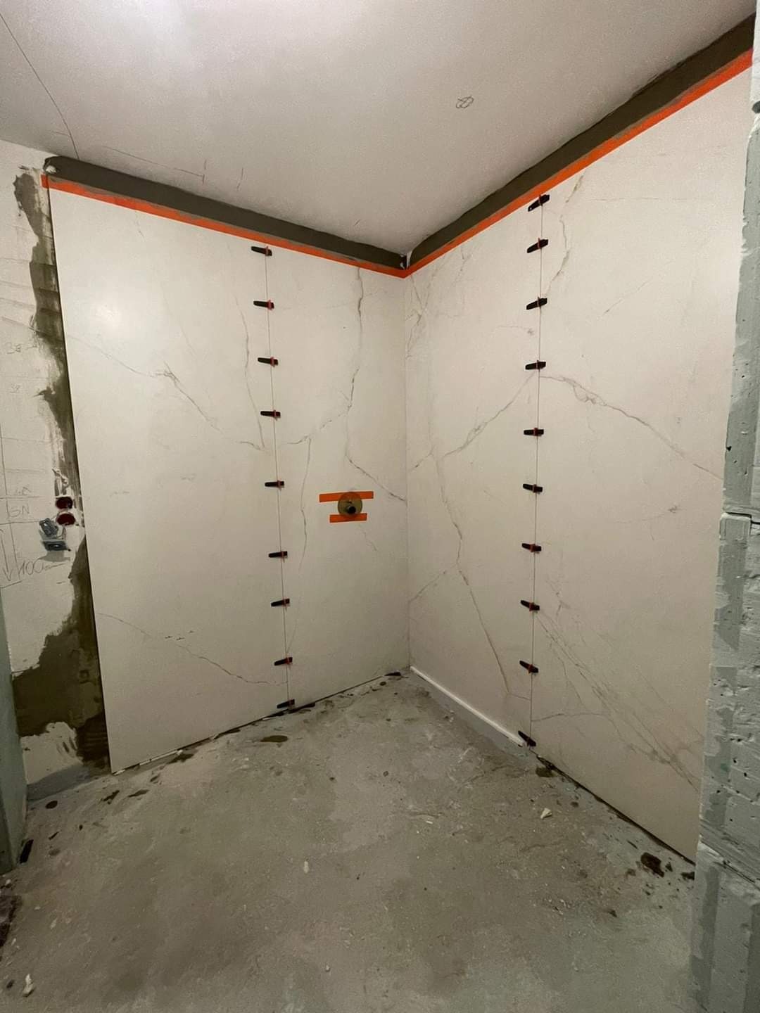 Wnętrze łazienki w trakcie układania płytek imitujących marmur na ścianach, widoczne systemy poziomowania płytek i surowa podłoga.