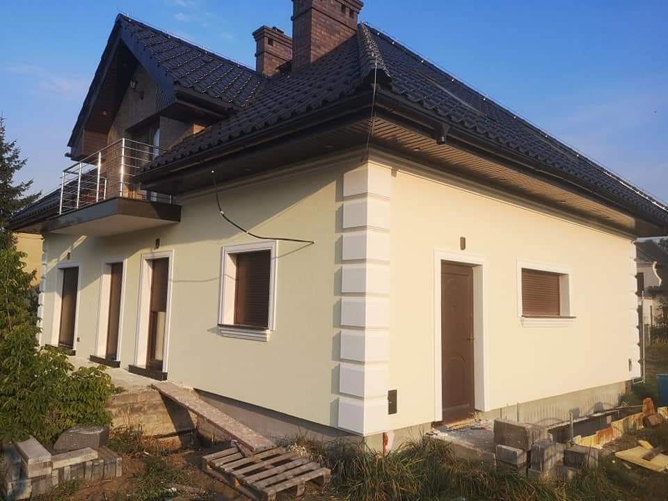 Jasnożółty dom jednorodzinny z ciemną dachówką i białymi narożnikami elewacji, widok z zewnątrz w słoneczny dzień.