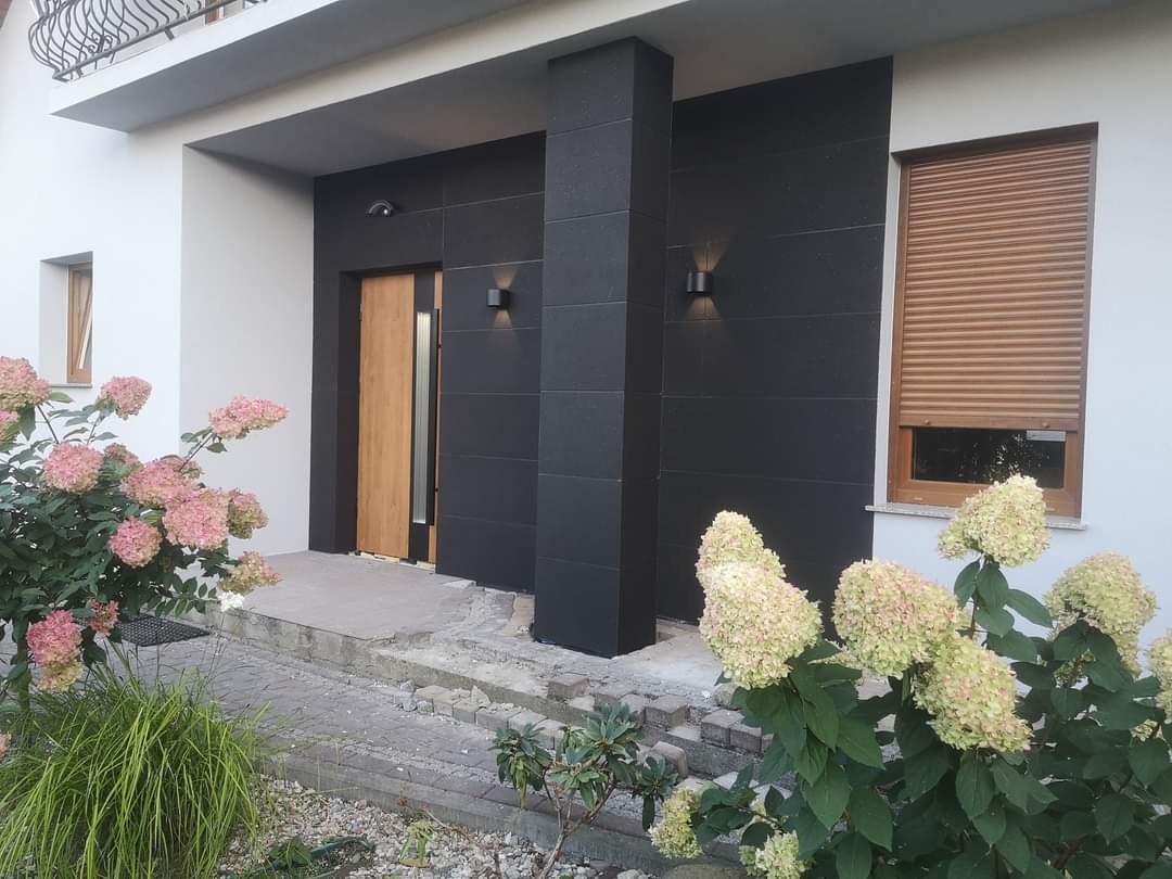Nowoczesny dom z czarną elewacją z płytek, drewnianymi oknami i drzwiami oraz oświetleniem zewnętrznym, otoczony kwitnącymi hortensjami, w trakcie prac wykończeniowych przy wejściu.