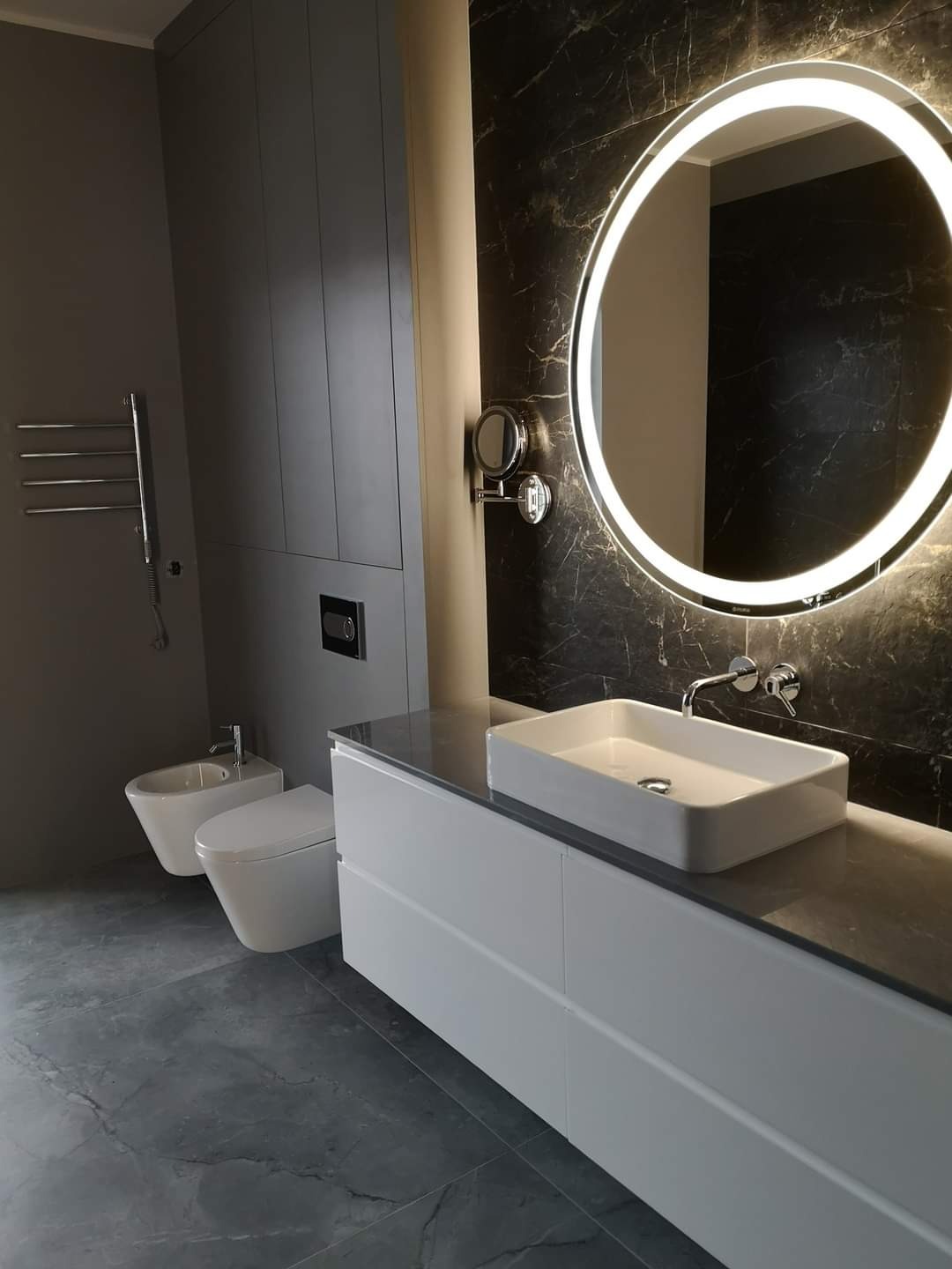 Nowoczesna łazienka w odcieniach szarości z podświetlanym okrągłym lustrem, białą umywalką nablatową i wiszącą toaletą oraz bidetem.