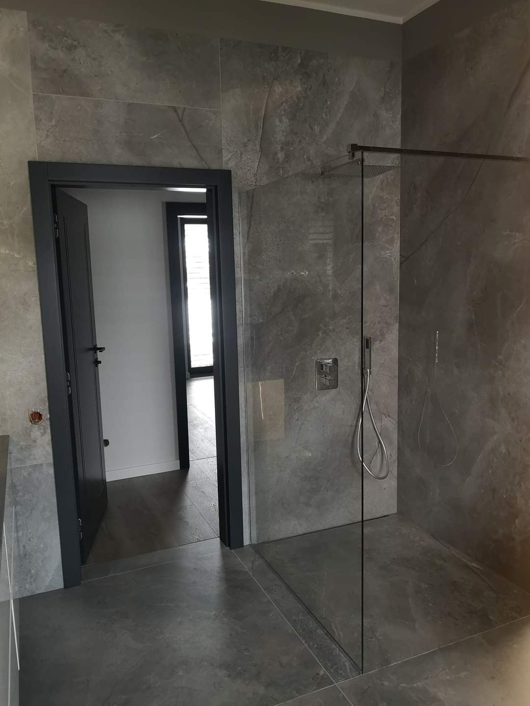 Nowoczesna łazienka z szarą, marmurkową okładziną ścienną i podłogową, kabina prysznicowa typu walk-in z chromowaną armaturą i szklaną ścianą, widok na otwarte drzwi do innego pomieszczenia.